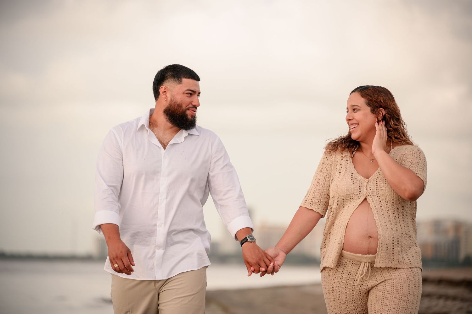 Pregnant couple holding hands and smiling at each other on a beach during a maternity photoshoot.