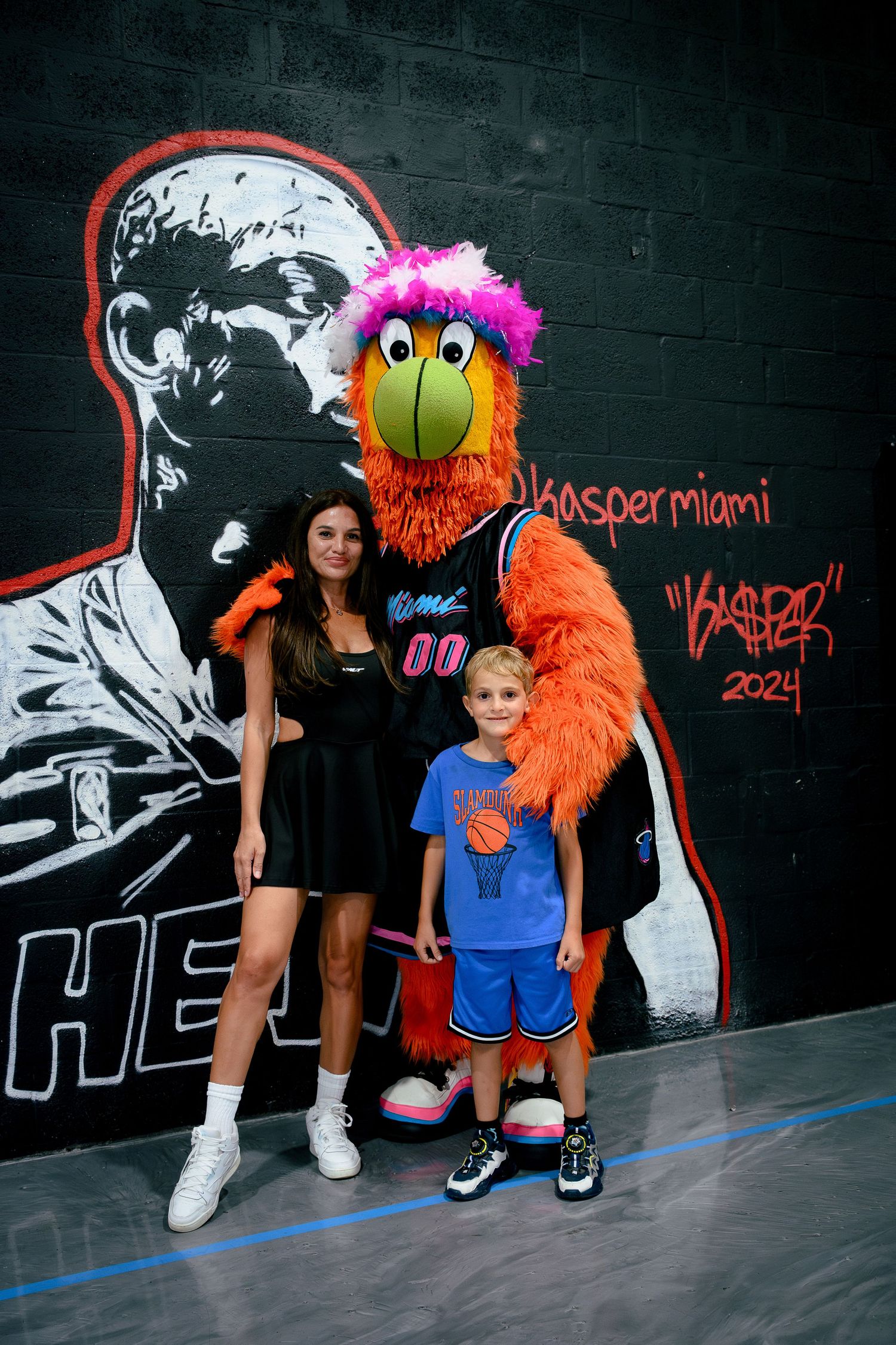 Woman and child pose with Miami Heat mascot Burnie in front of a graffiti mural at a basketball court.