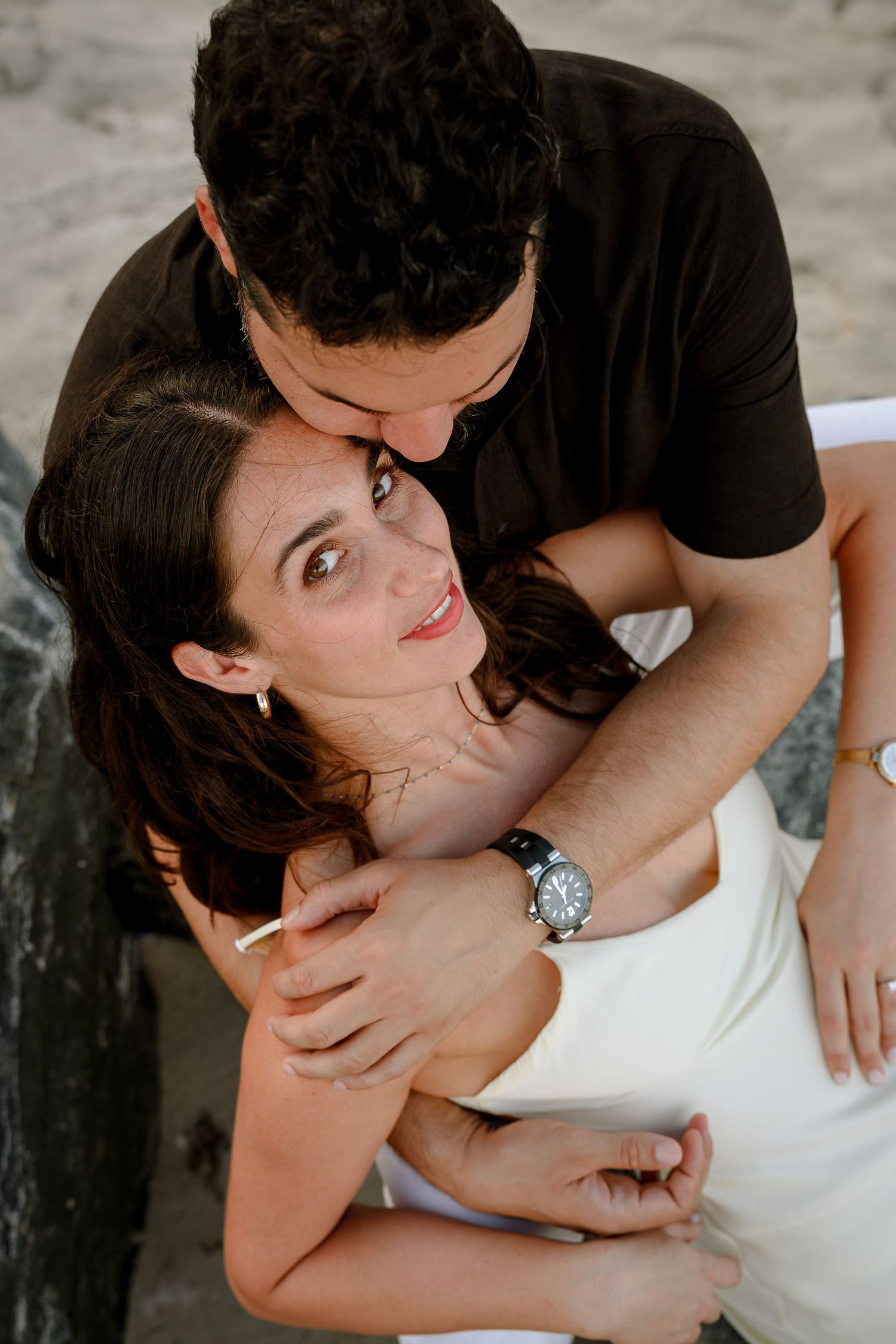A romantic couple embraces outdoors, woman in white smiles up as man in black kisses her forehead, wearing a watch.