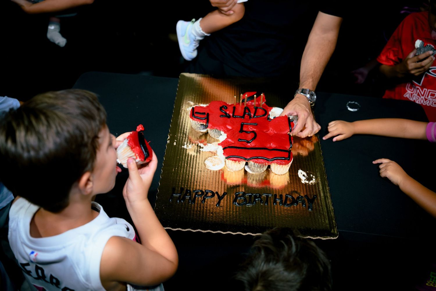 Children gathered around a red Happy Birthday cake with number 5 candle at a birthday party celebration.