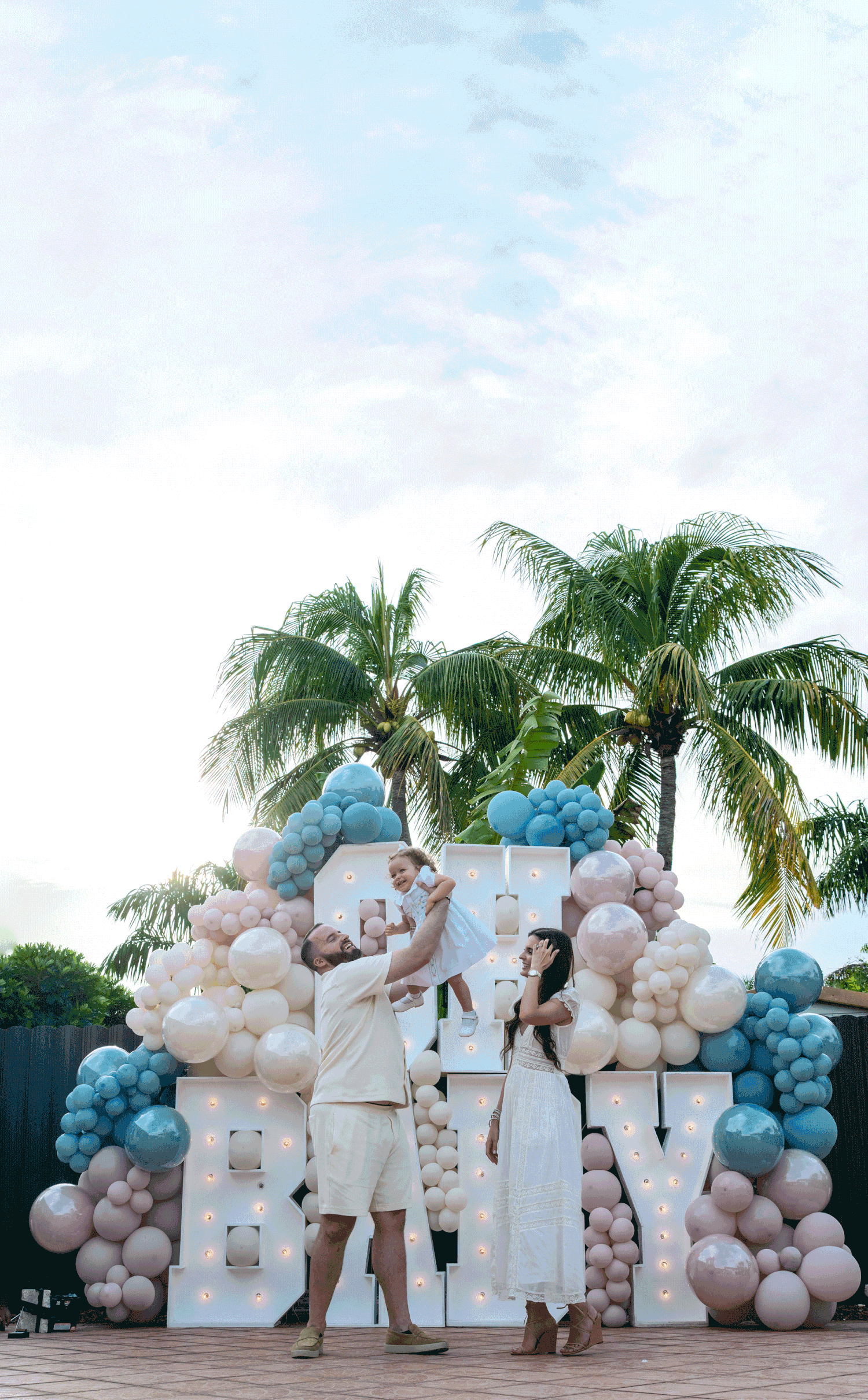 Large white balloon letters spelling 'BABY' with blue and white balloon arch decorations against palm trees outdoors.