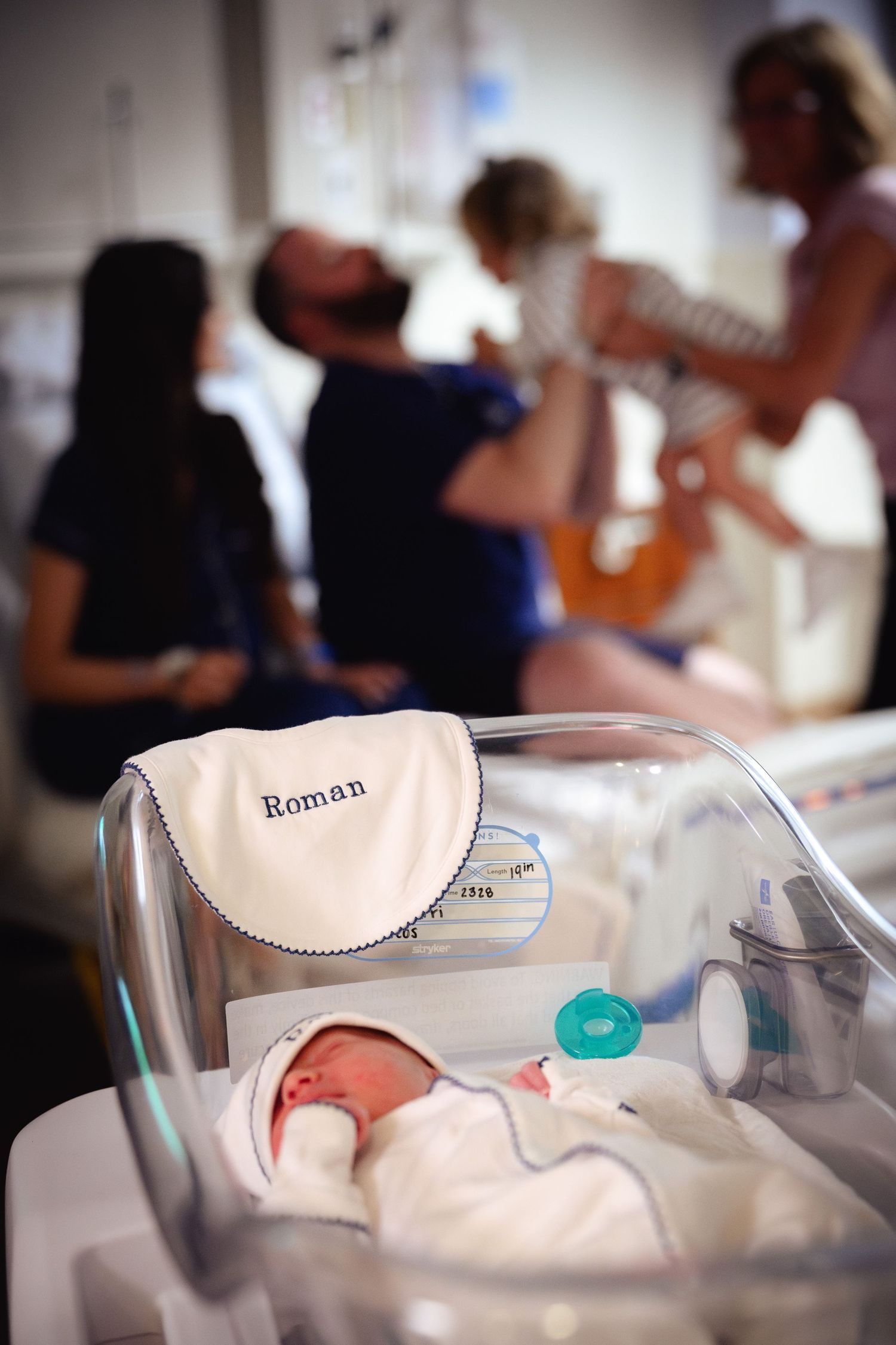 A newborn baby named Roman rests in a hospital bassinet with a pacifier while family gathers in the background.