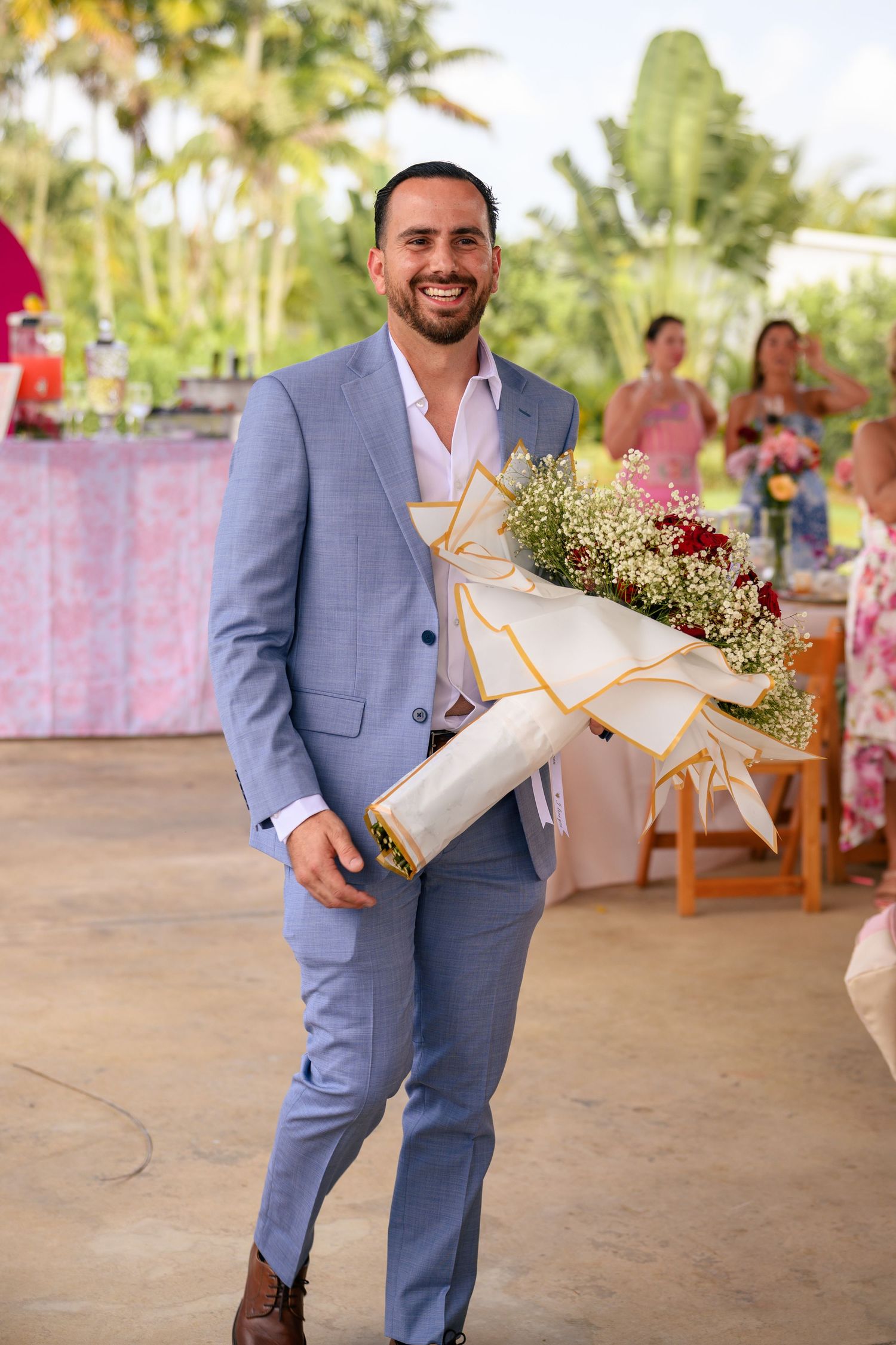 A person in a light blue suit holds an elegantly wrapped bouquet at an outdoor event venue.