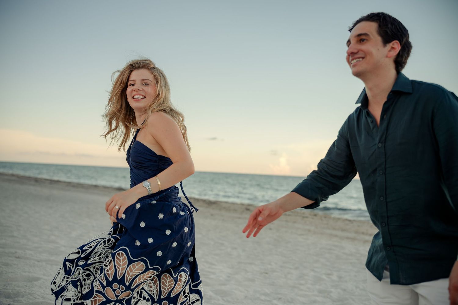 Happy couple laughing and running together on a sandy beach at sunset, woman in blue floral dress.