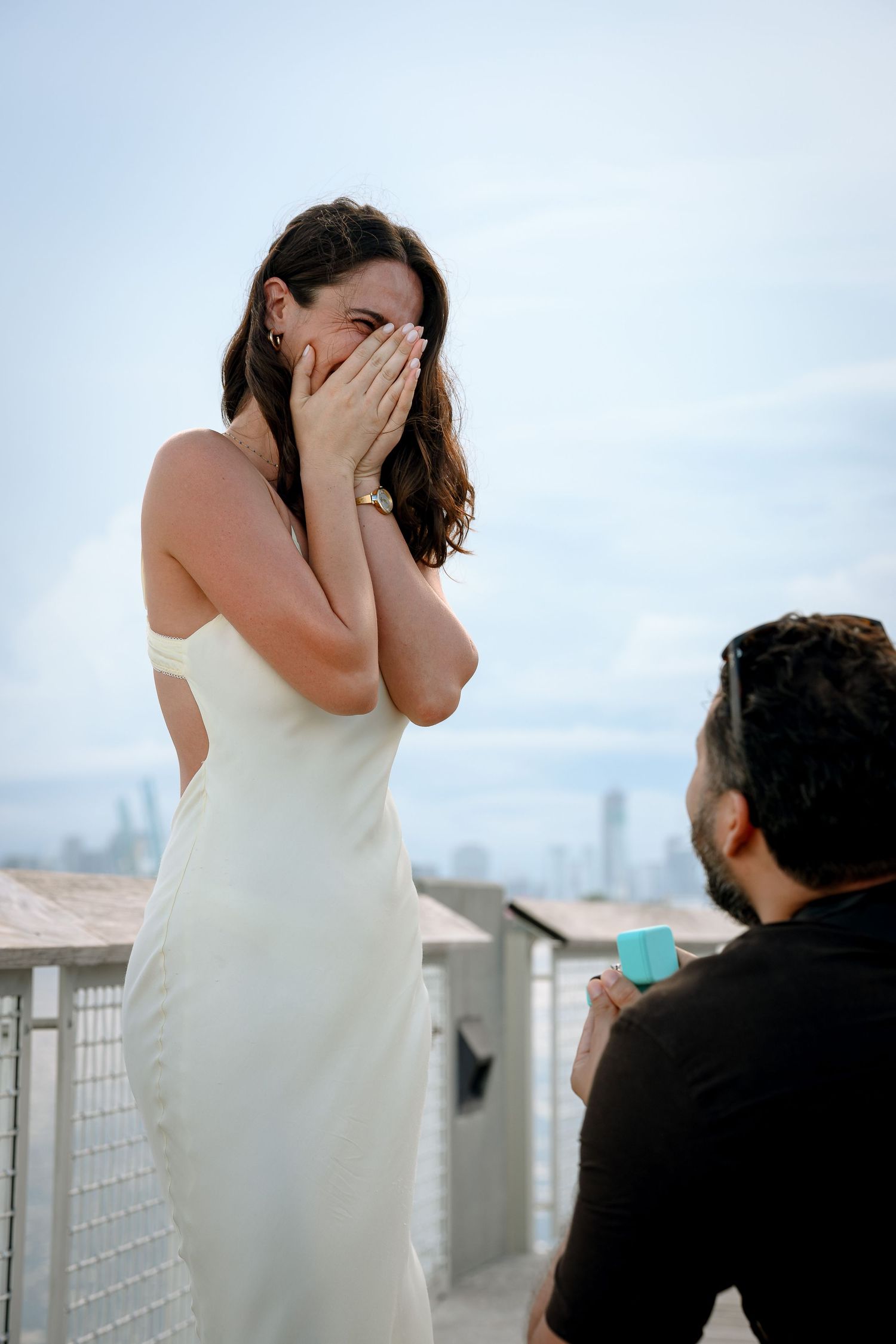 A man kneeling to propose to a surprised woman in a white dress on a rooftop with a city skyline backdrop.