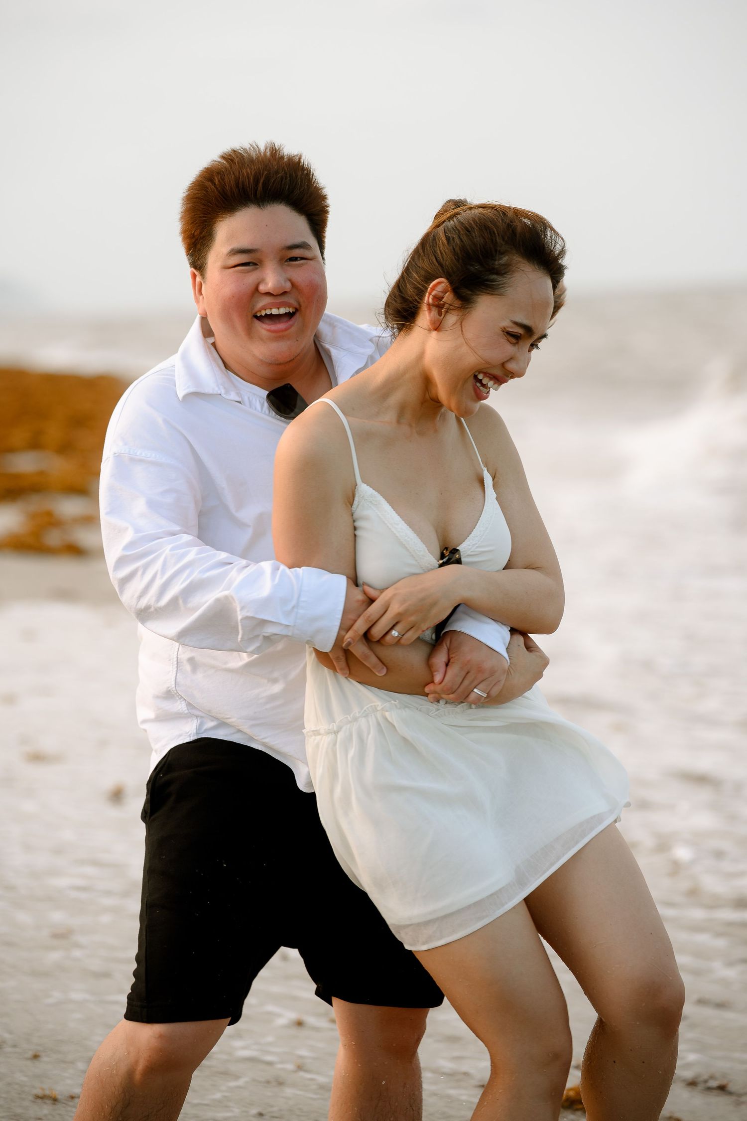 Couple laughing joyfully on a beach, woman in white dress and man in white shirt embracing by the ocean waves.