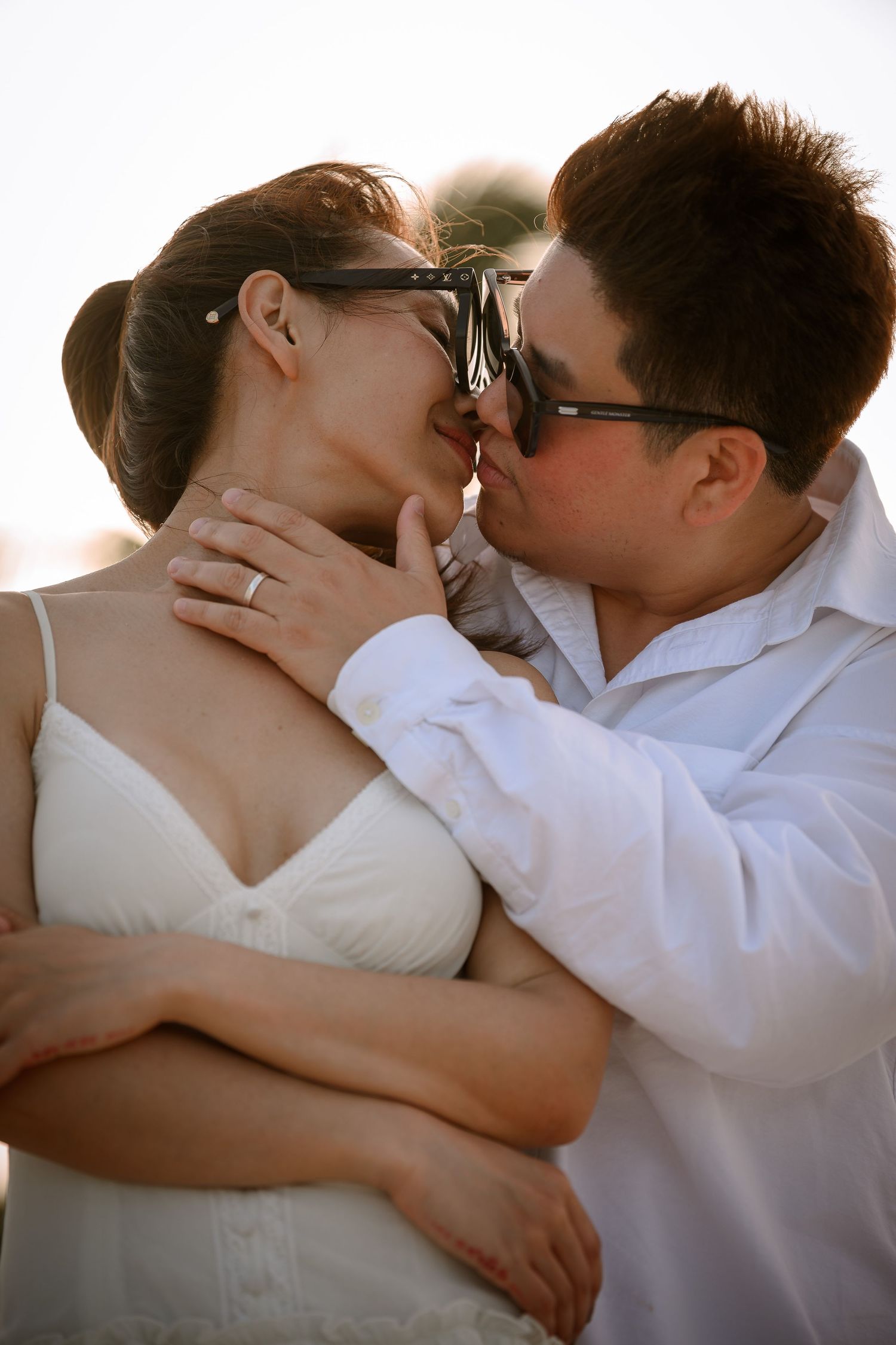 Couple in sunglasses sharing a romantic kiss, woman in white dress with engagement ring, man in white shirt.
