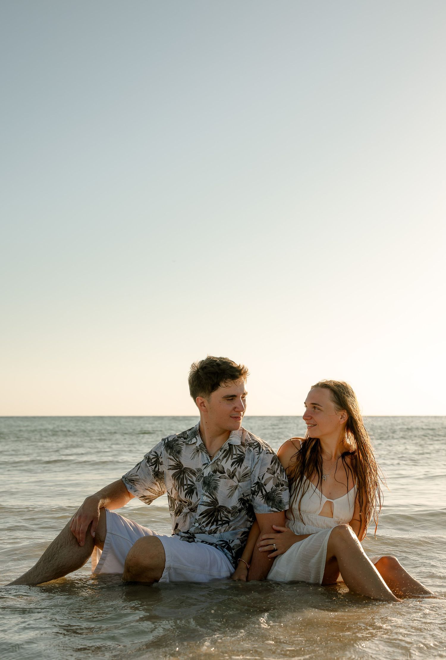A couple sits together in shallow ocean water at sunset, smiling at each other in casual summer attire.