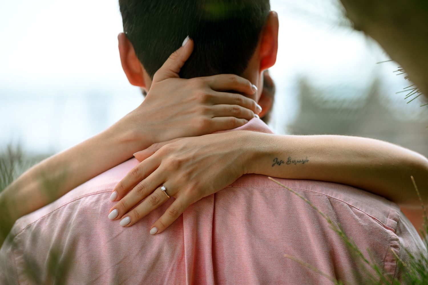 Couple embracing from behind, woman's arm with tattoo around man's neck, wearing pink shirt outdoors.
