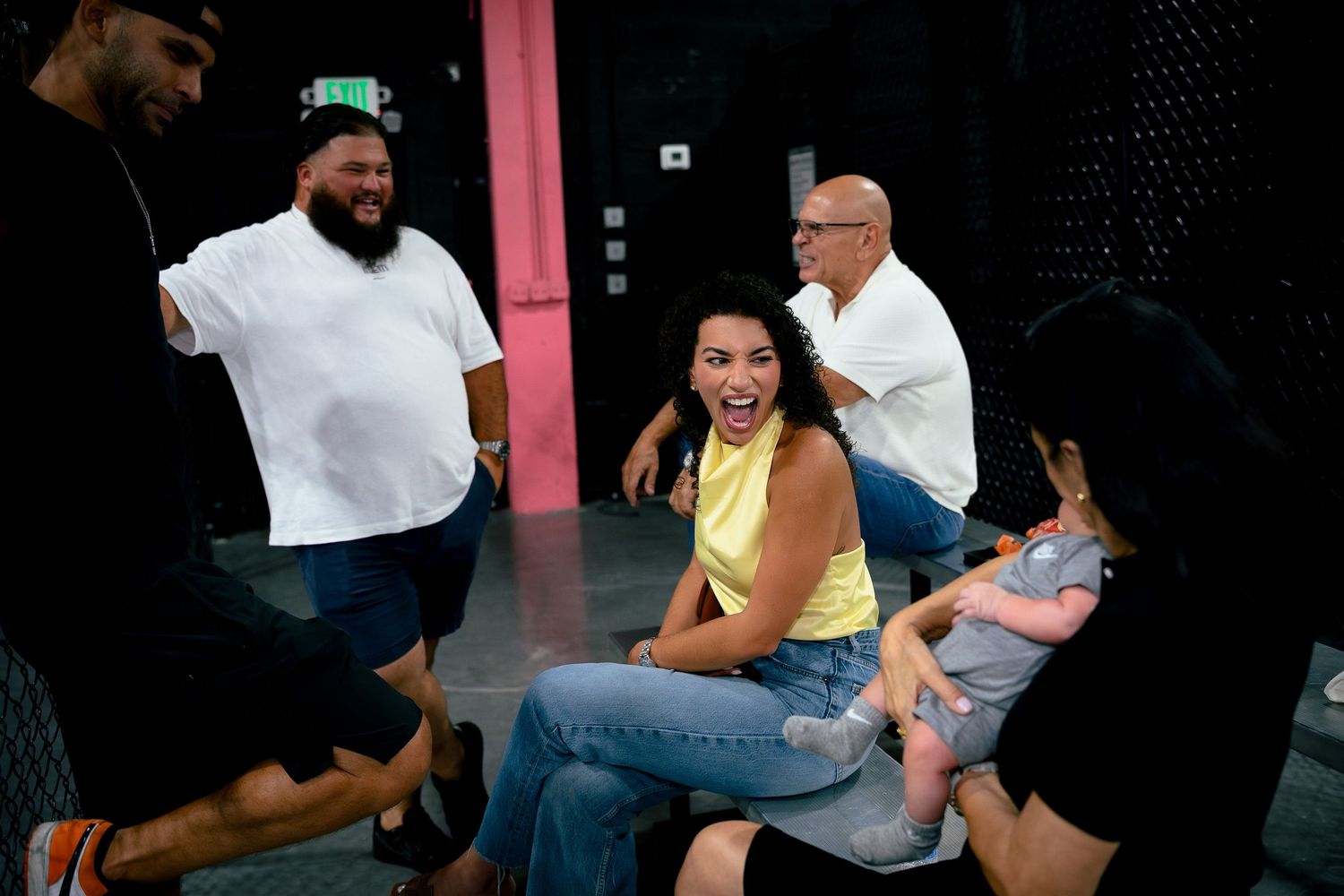 A group of diverse people laughing and socializing outdoors, with a woman in yellow holding a baby.