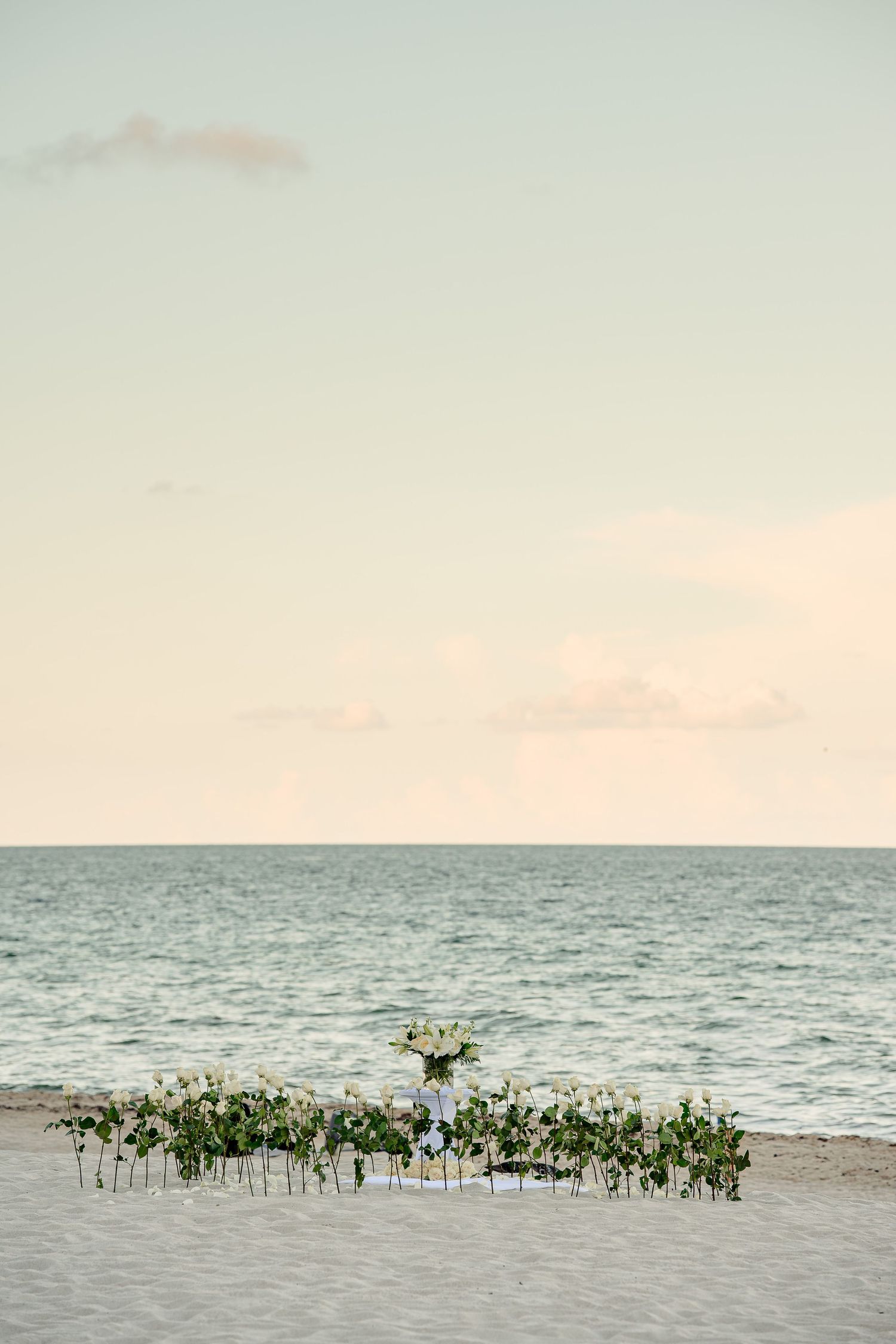 Elegant beach wedding setup with white floral arrangements on golden sand, overlooking a calm turquoise ocean at sunset.