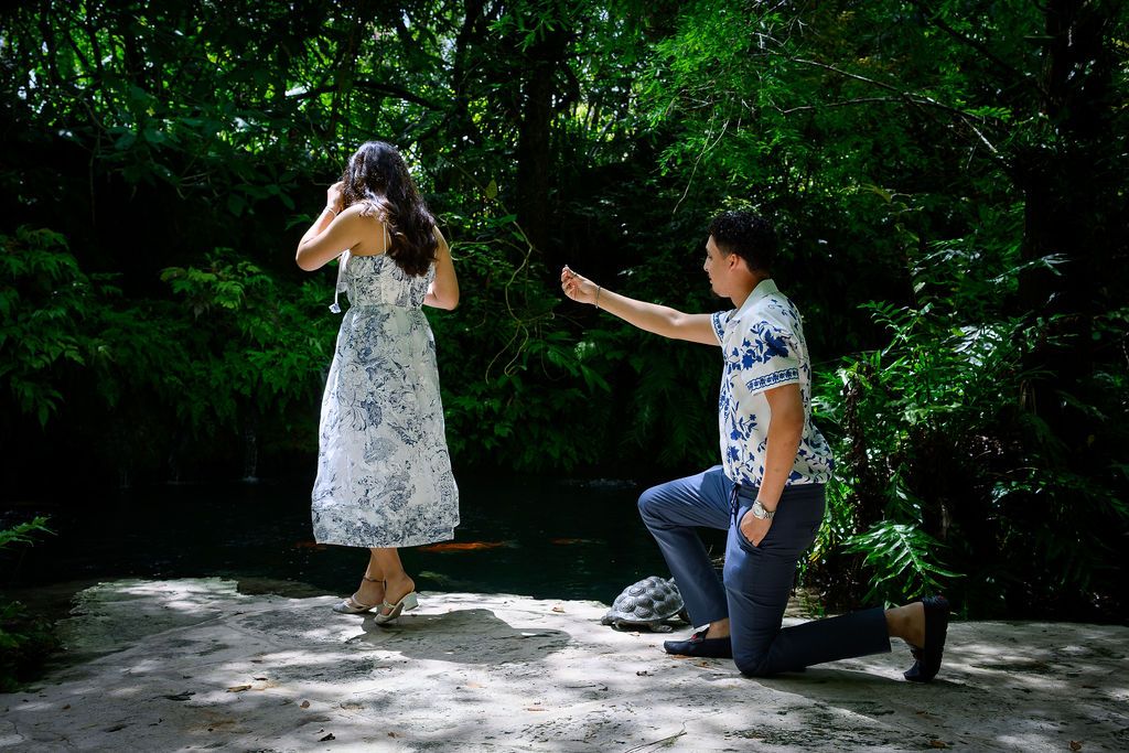 Man kneeling on one knee proposing to a woman in a lush green forest setting near a serene pond.