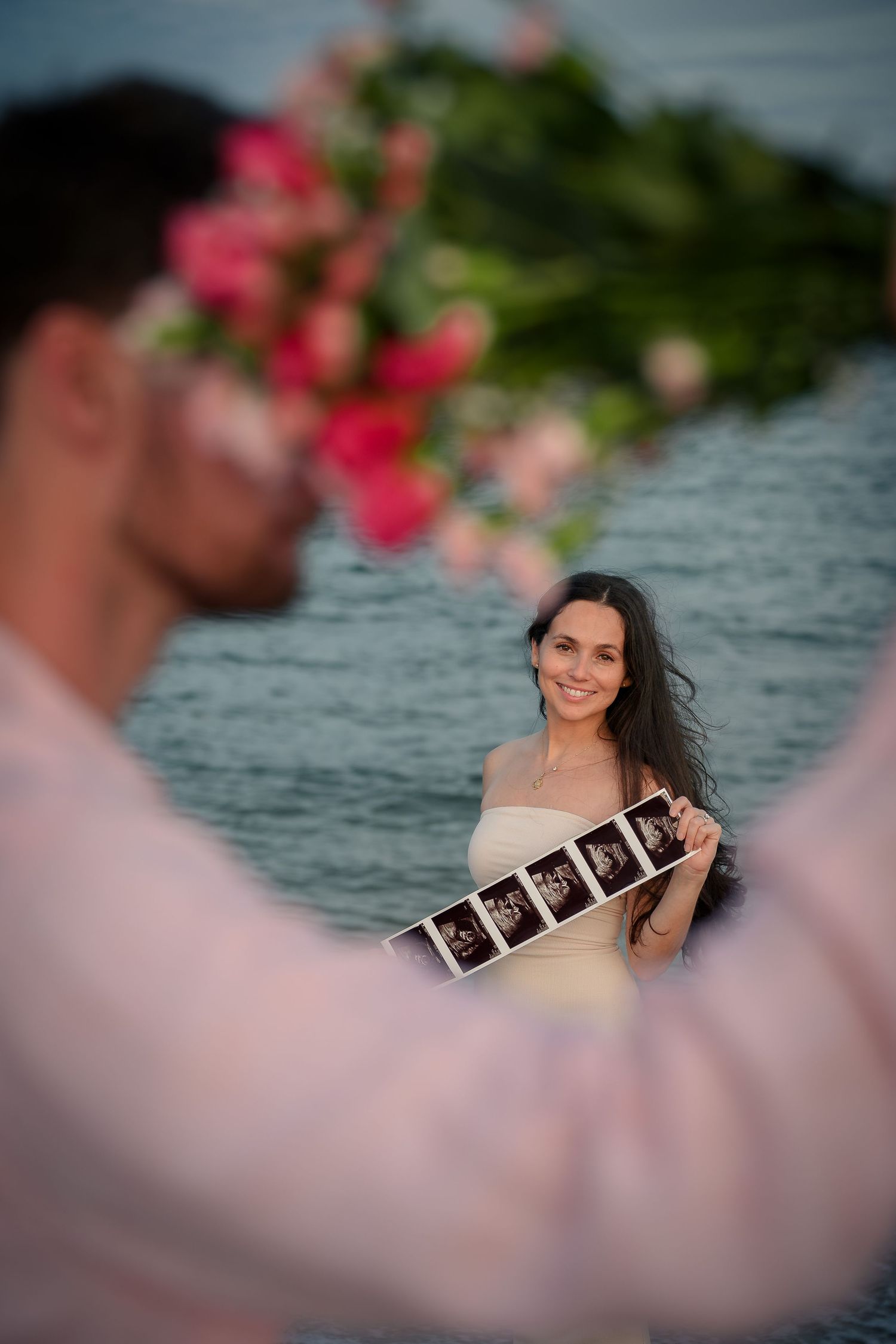 Smiling woman holds ultrasound photos near water with pink flowers, sharing pregnancy news with partner outdoors.