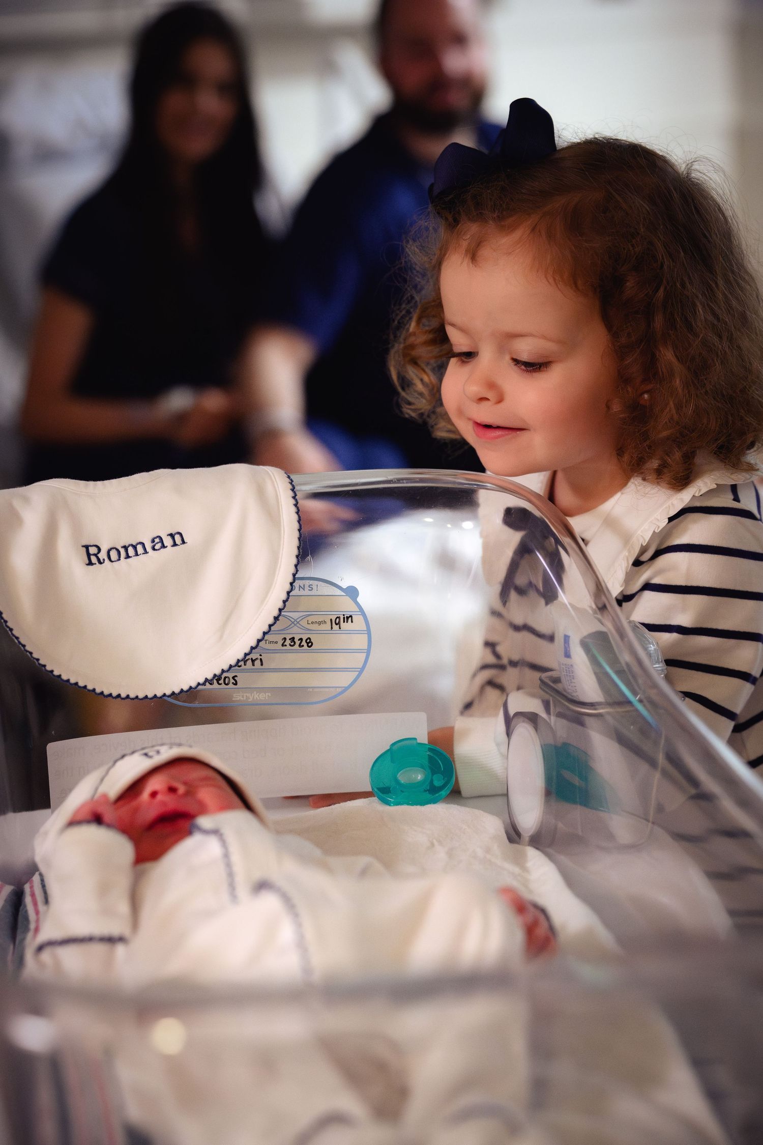A child in a striped shirt looks at a newborn baby in a hospital bassinet labeled 'Roman'.