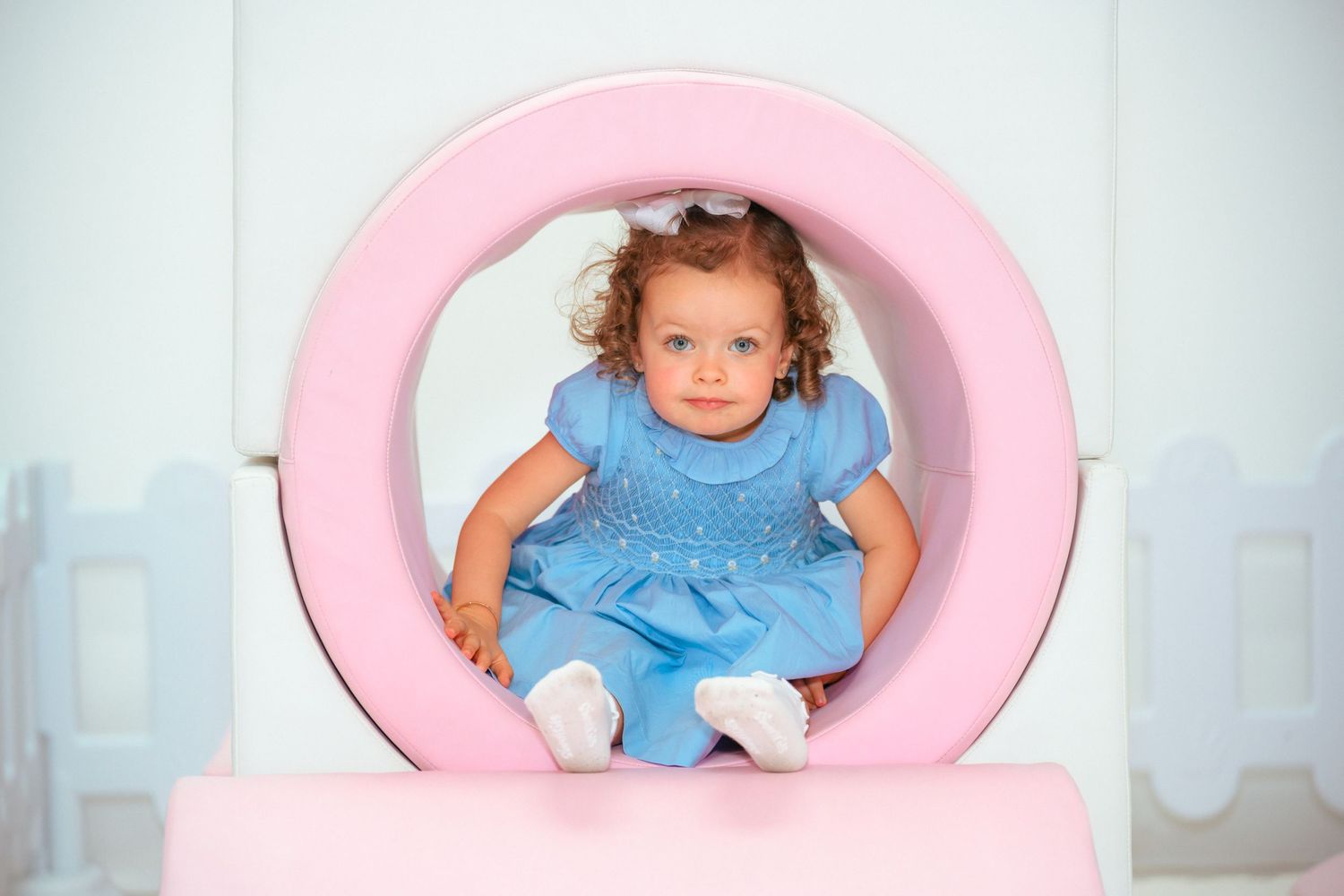 Curly-haired toddler girl in blue dress sitting inside a pink circular frame at a children's play area.