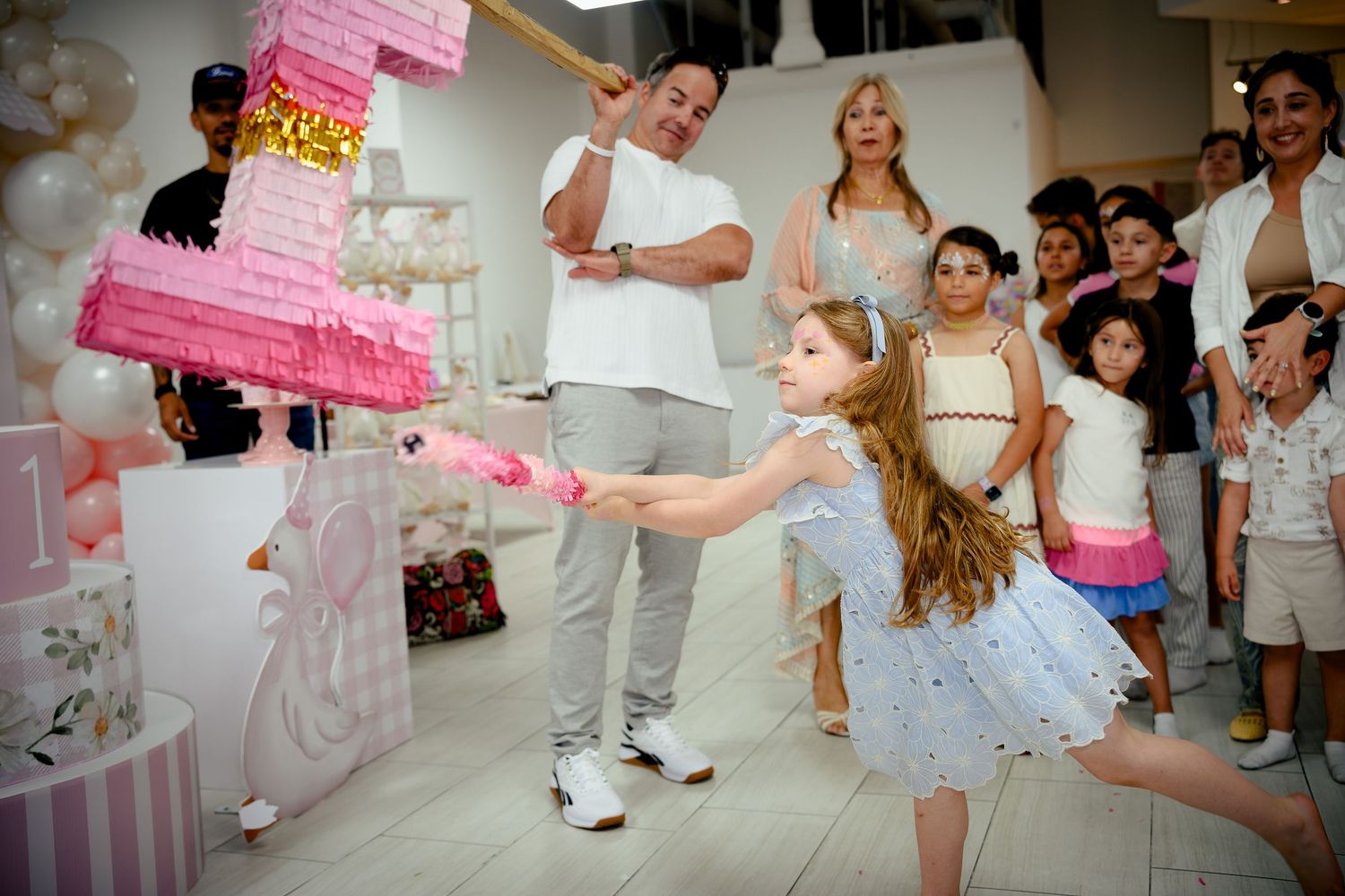 Young girl hitting a pink pinata at a birthday party while adults watch and cheer indoors.