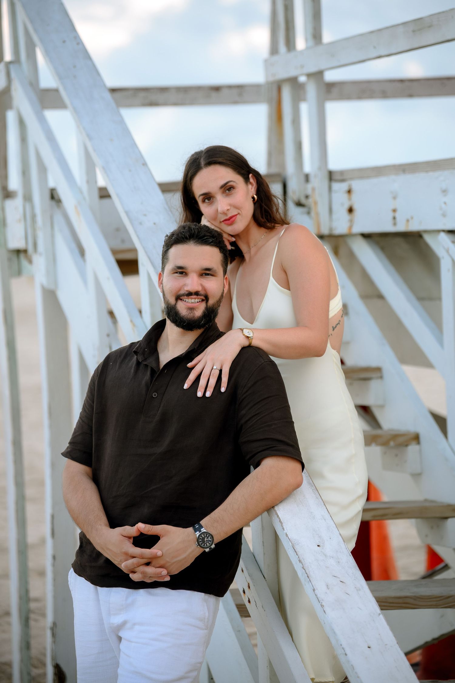 Couple posing on white wooden beach stairs, man in black polo and woman in white dress, smiling at camera.