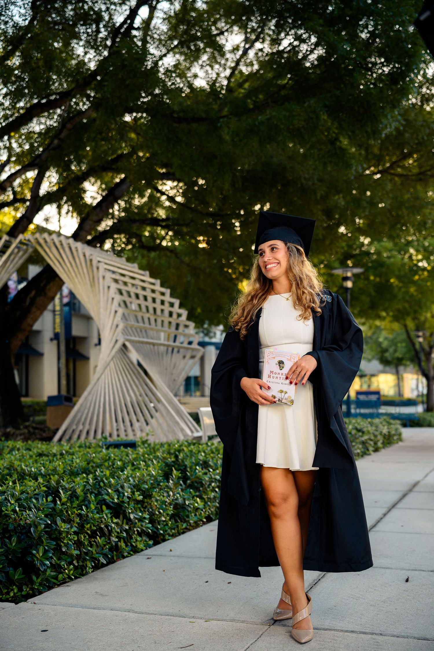 Smiling female graduate in black cap and gown holding diploma while walking on sunny campus pathway.