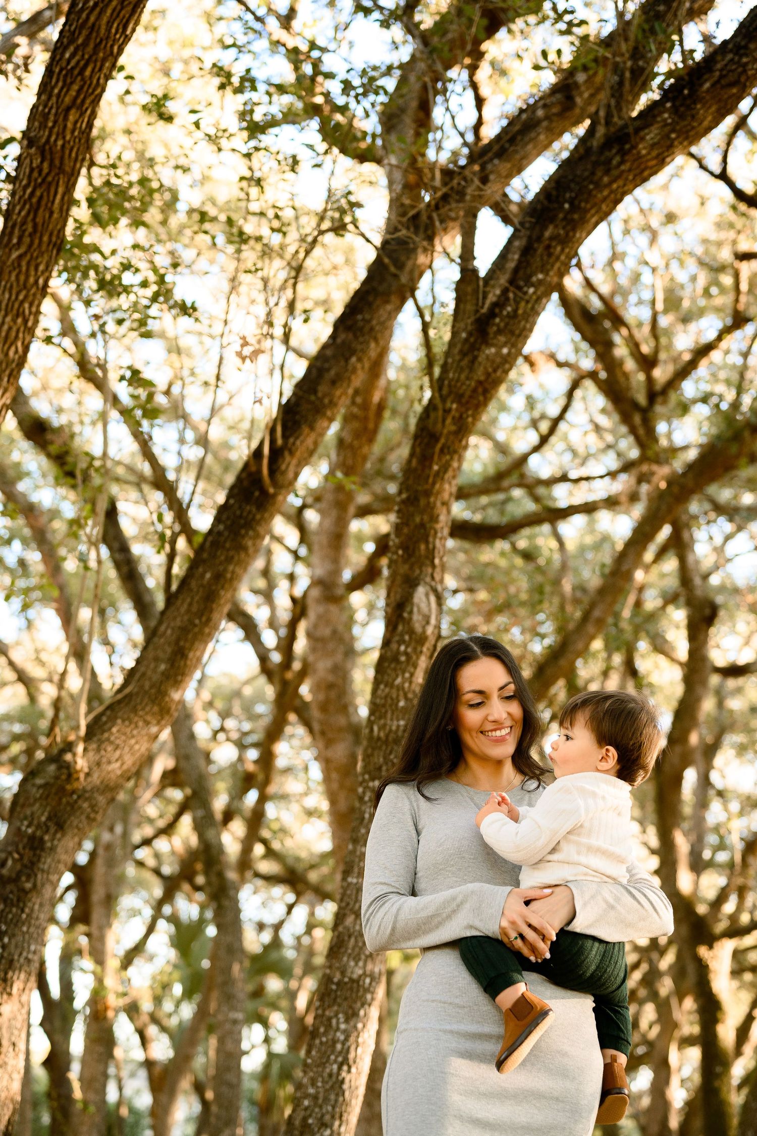 Smiling mother holds baby while standing under golden sunlit oak trees in park.