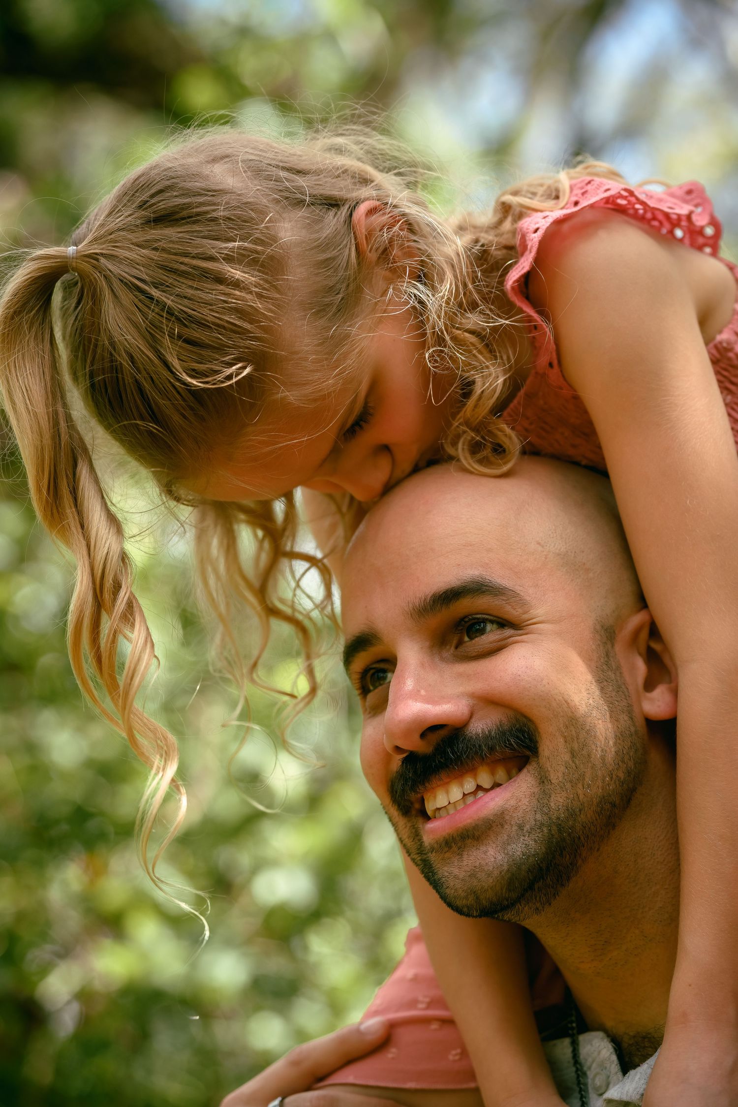 A smiling father with a beard carries his young curly-haired daughter on his shoulders outdoors.