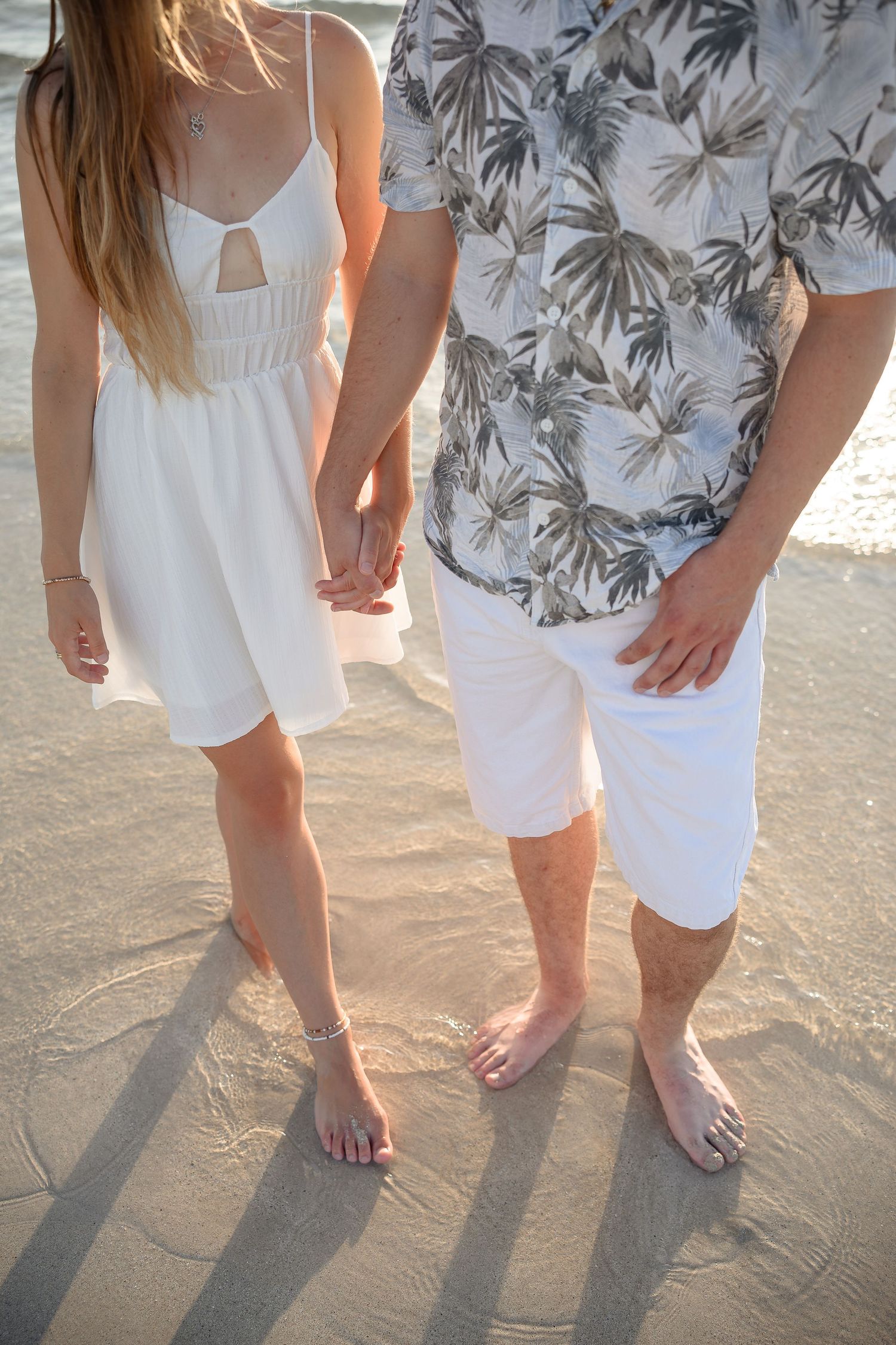 Couple holding hands barefoot on sandy beach, woman in white dress and man in tropical shirt with white shorts.