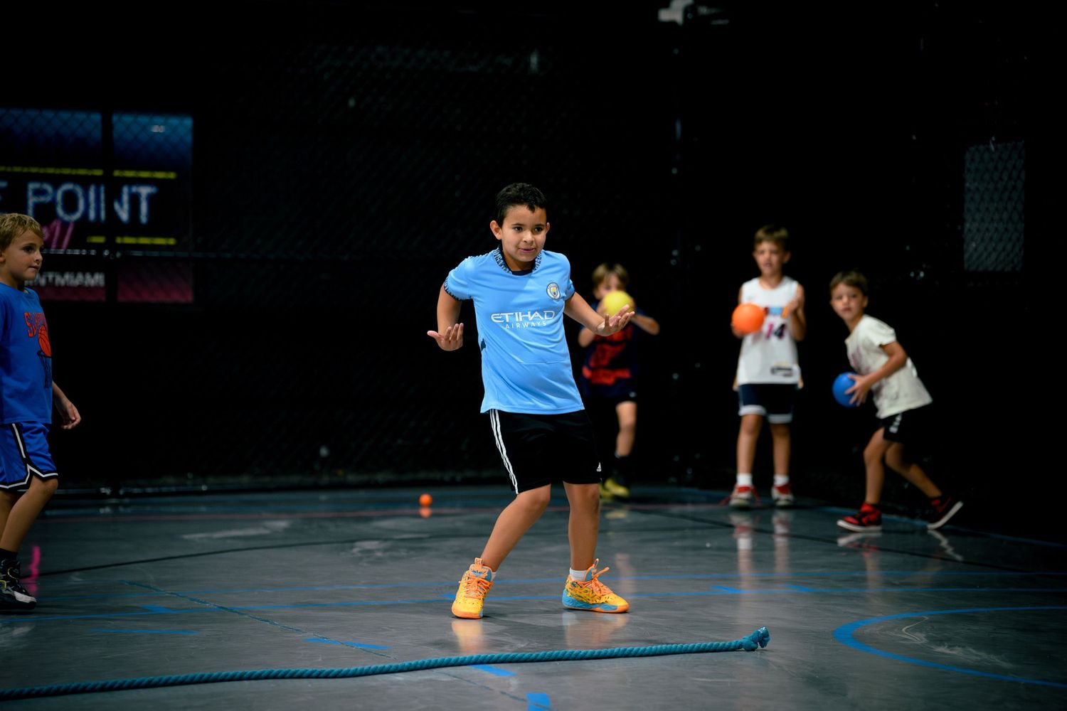 Children playing dodgeball in a gym, with a boy in a blue shirt mid-game on a marked court.