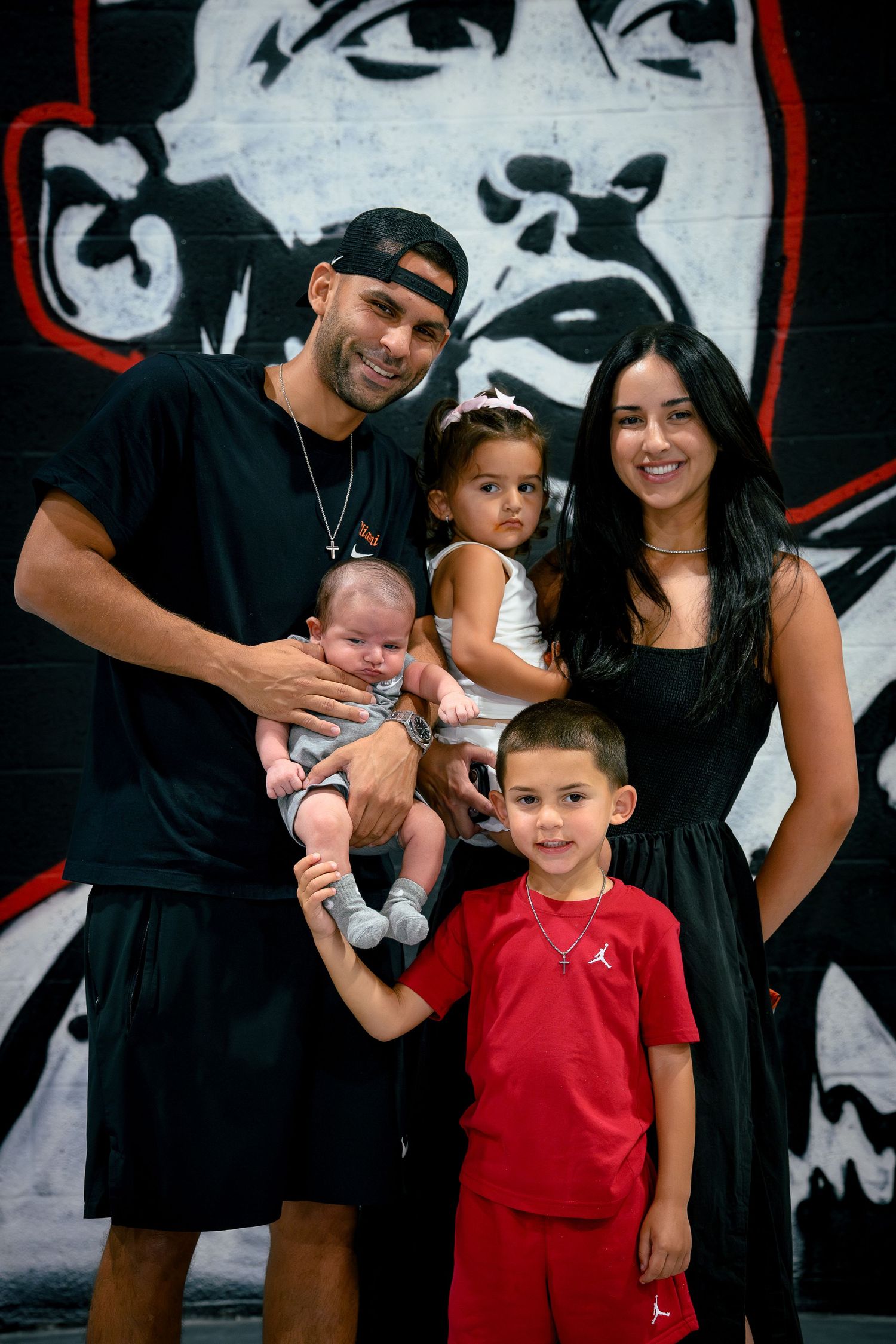 A family of five posing together in front of a bulldog mural, including parents holding a baby, a toddler girl, and a young boy.