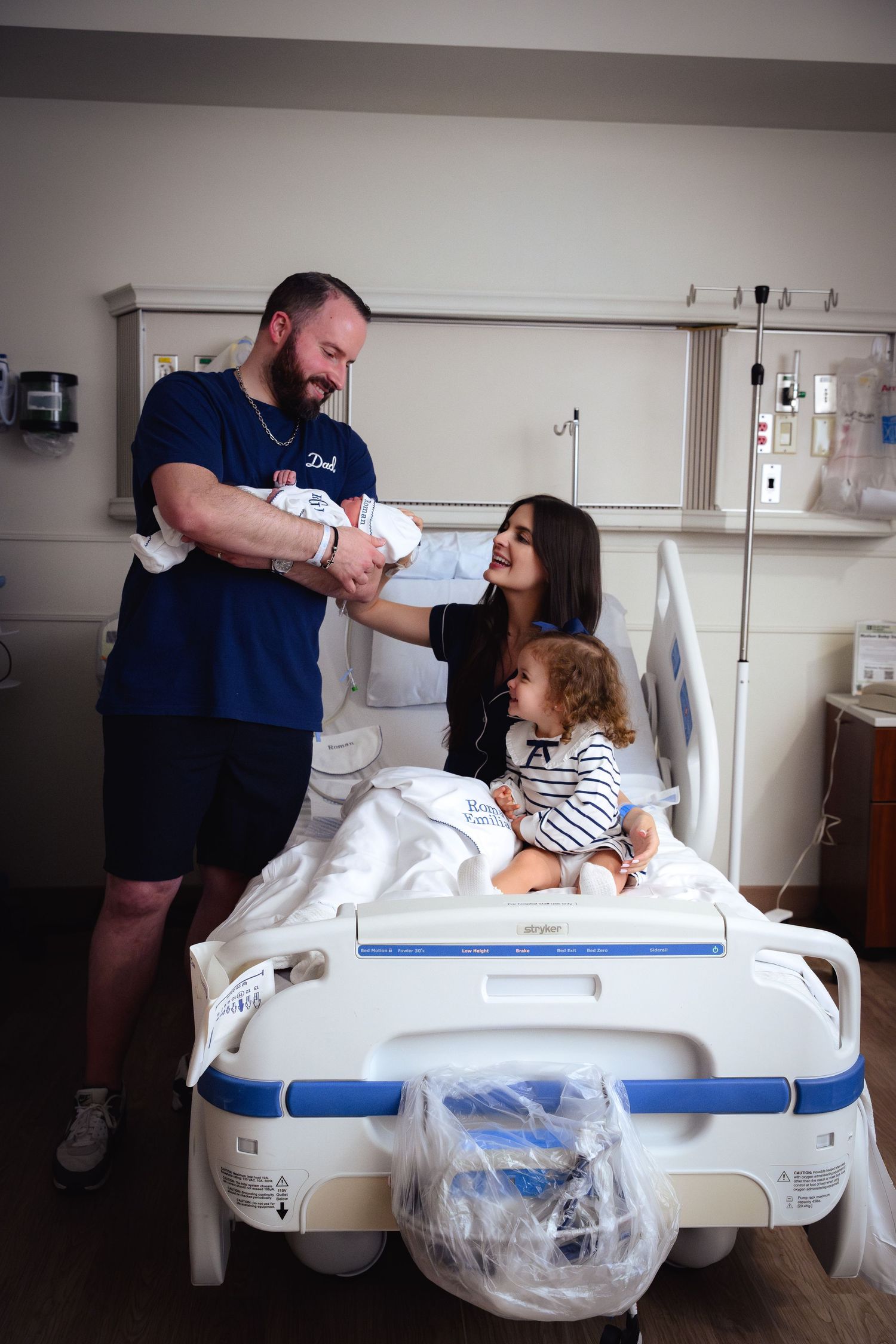 Family celebrating new baby in hospital room with dad, toddler, and mom on hospital bed.