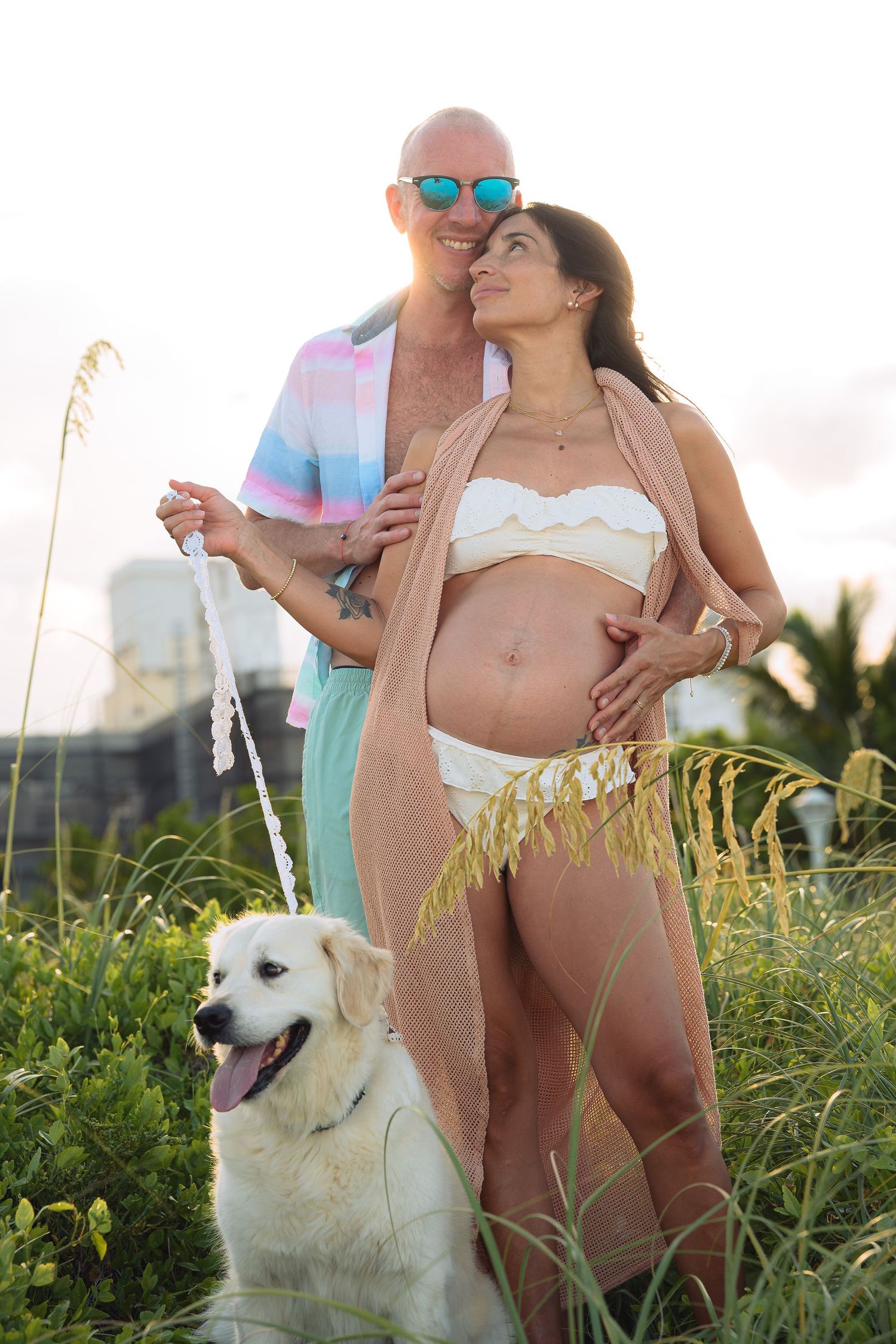 Pregnant woman in white bikini with partner and white dog posing outdoors in tall grass at sunset.
