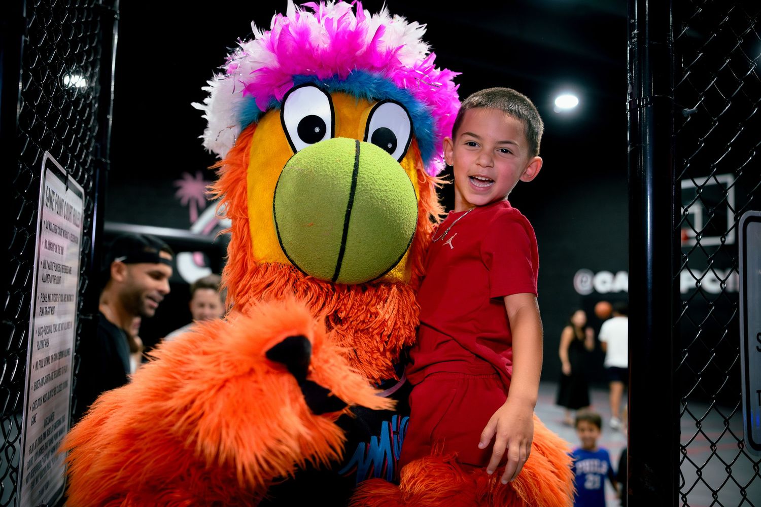 A smiling boy poses with a colorful orange and pink bird mascot inside a cage at a sports event.