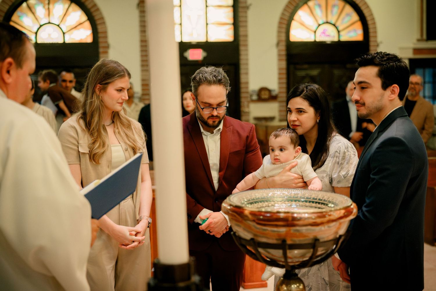 A baptism ceremony takes place in a church as family gathers around the baptismal font with a priest.