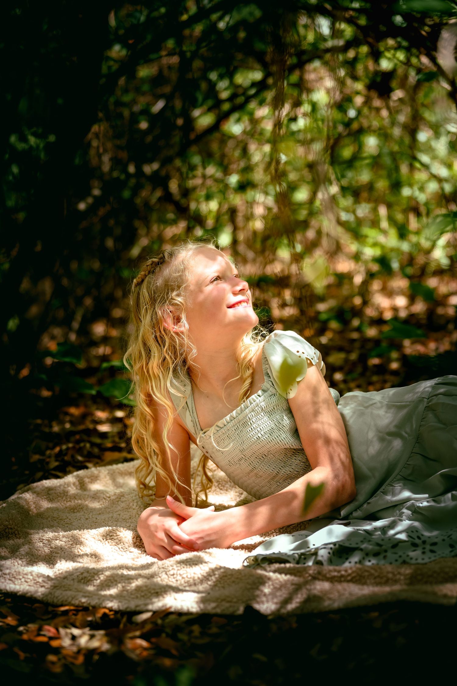 Young blonde girl in a white dress lying on a blanket in a sunlit forest, smiling and looking up.