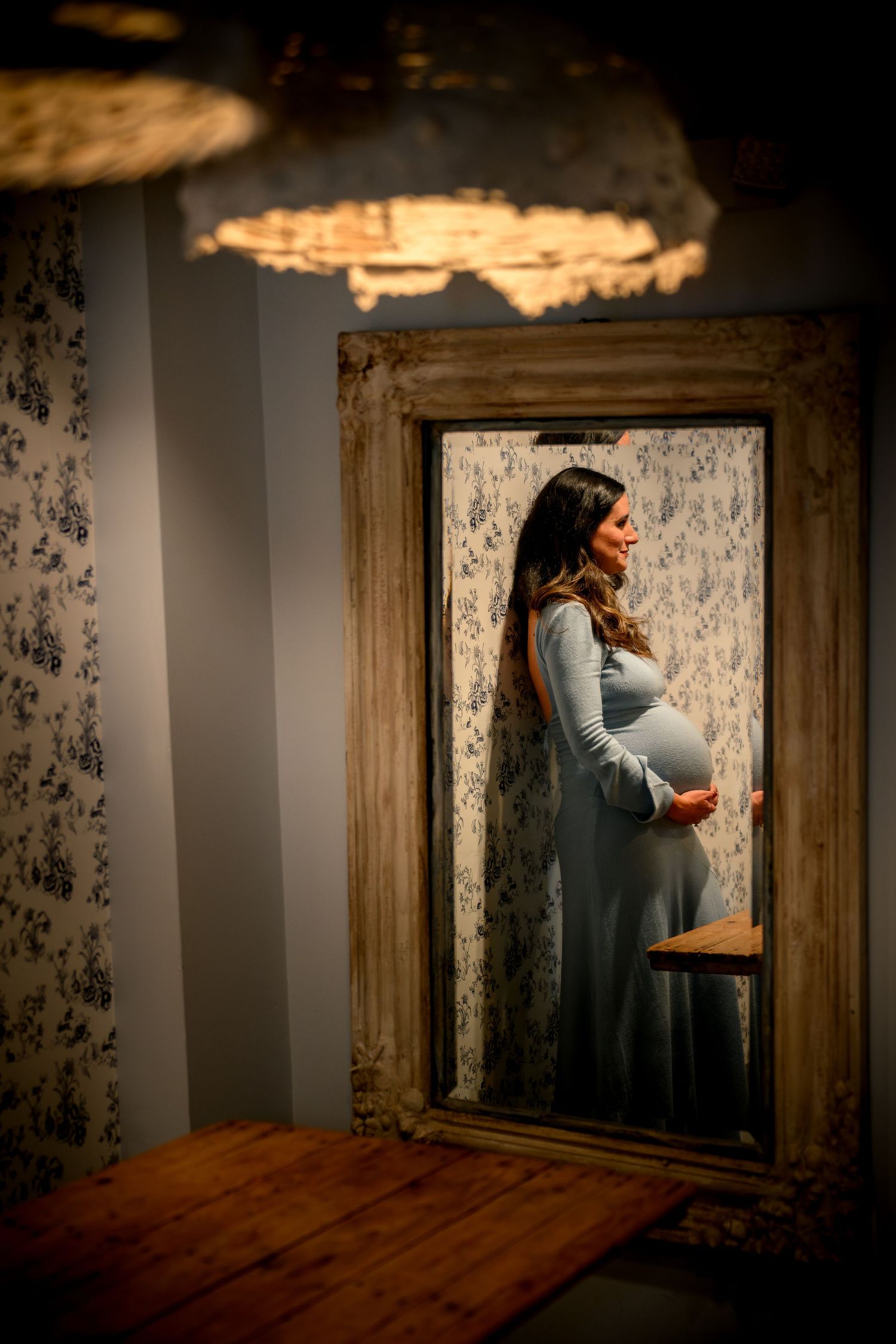 Moody maternity photoshoot in a rustic room with floral wallpaper and dramatic lighting.