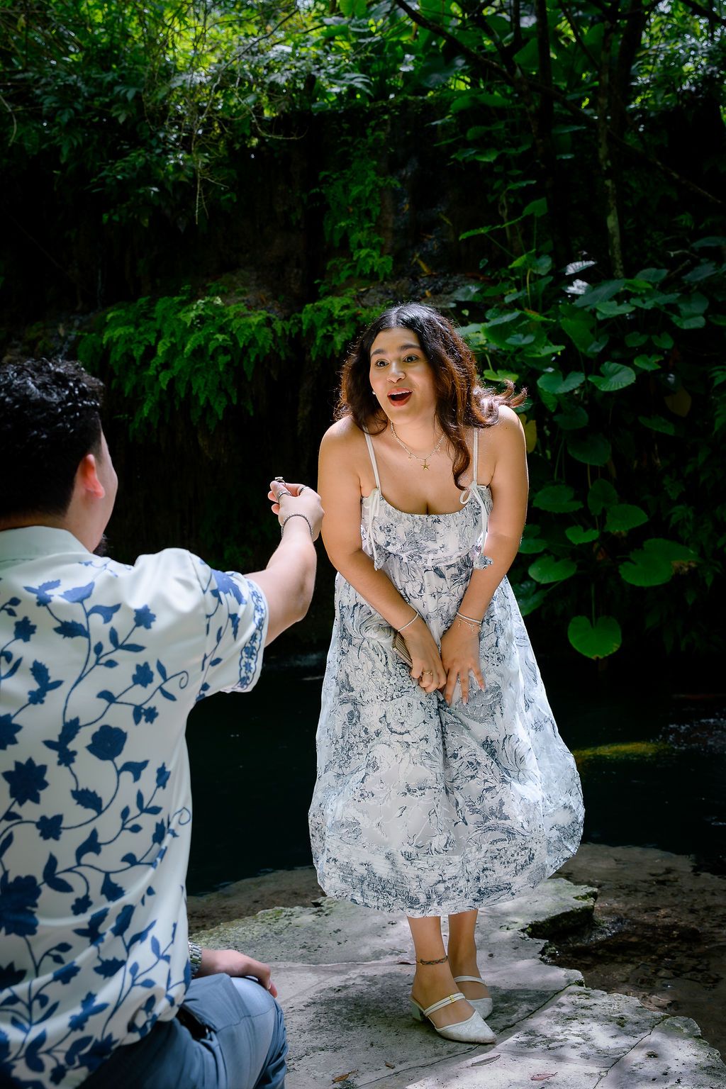 Man kneeling outdoors proposing to surprised woman in floral dress near lush green tropical garden.
