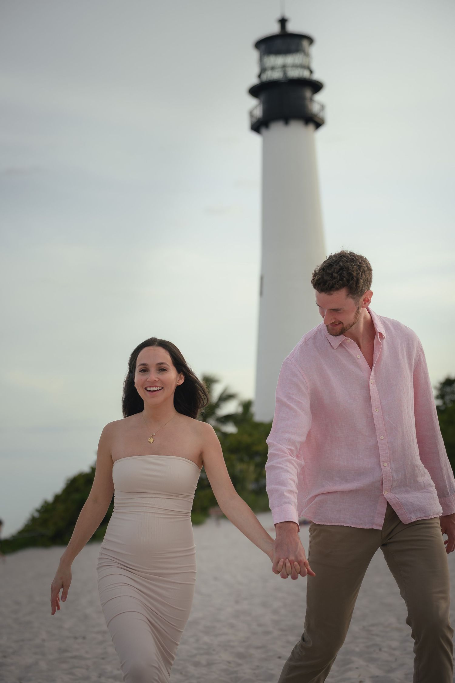 Couple holding hands walking on beach near lighthouse, woman in nude strapless dress, man in pink linen shirt.