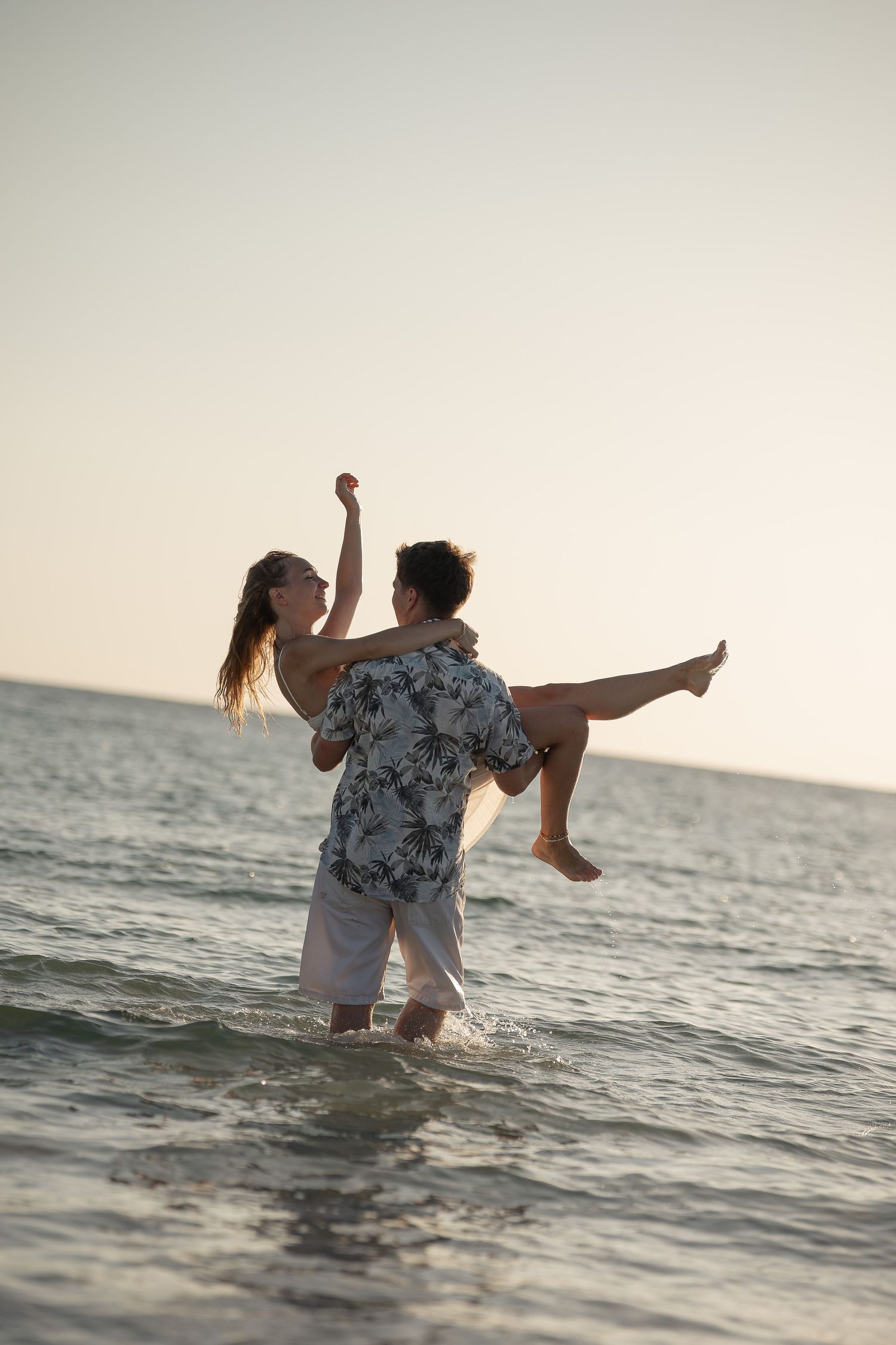 Couple playing in ocean waves at sunset, man lifting woman in his arms while she raises her fist joyfully.