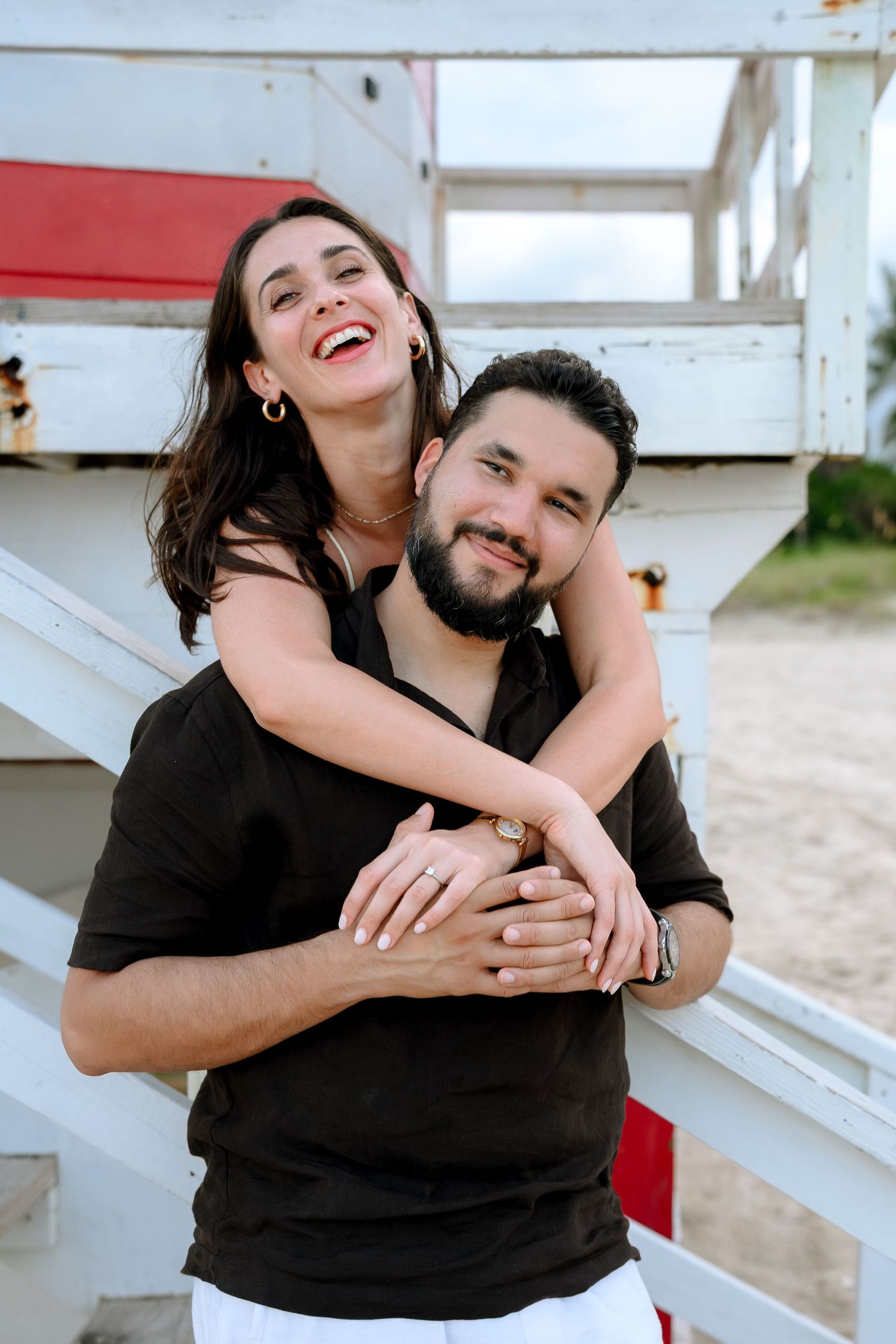 Happy couple in black outfits embracing near a beach lifeguard stand, woman laughing joyfully.