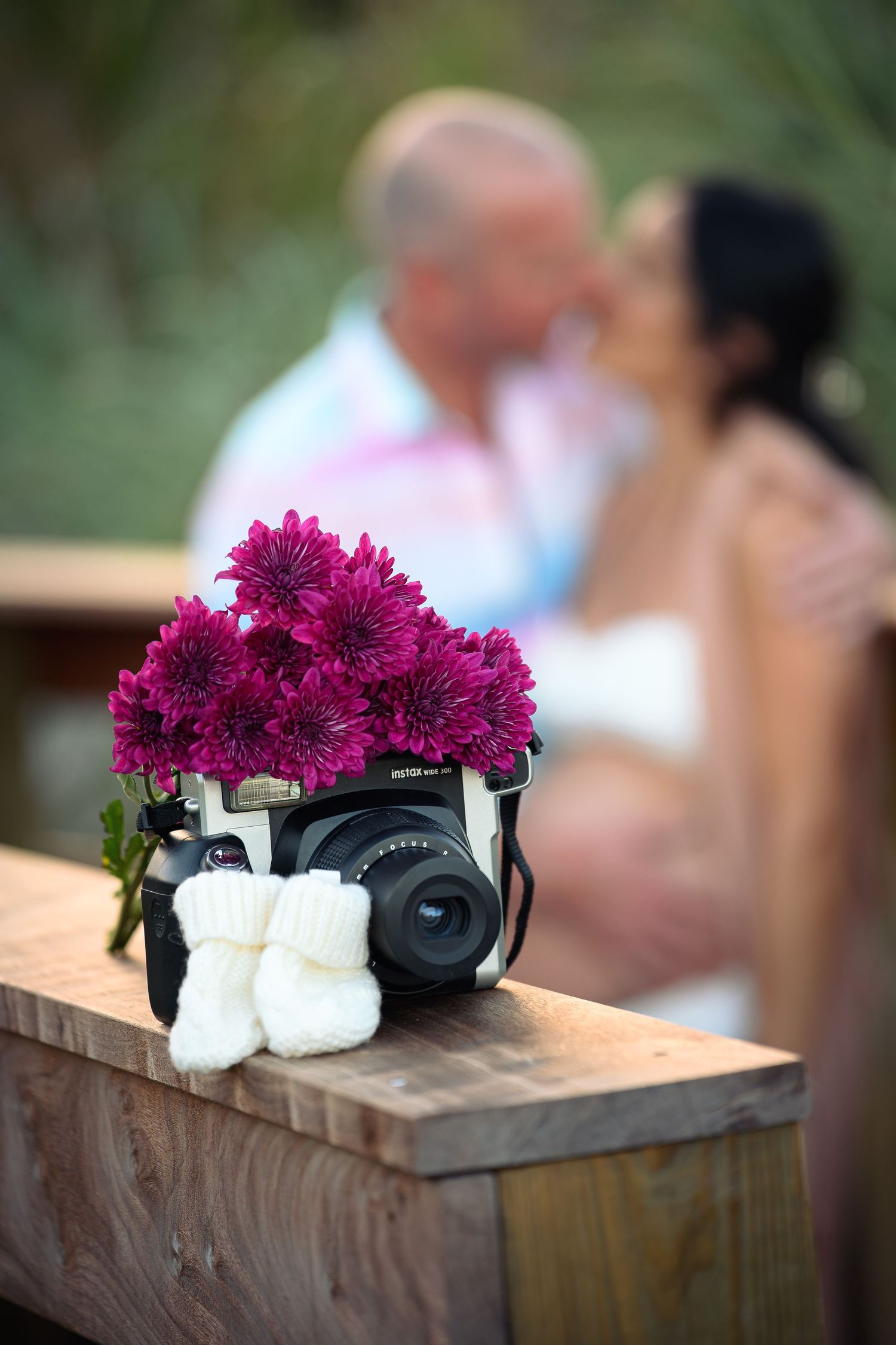 Fujifilm instax camera with purple chrysanthemums and white baby booties on wood, couple kissing in background.