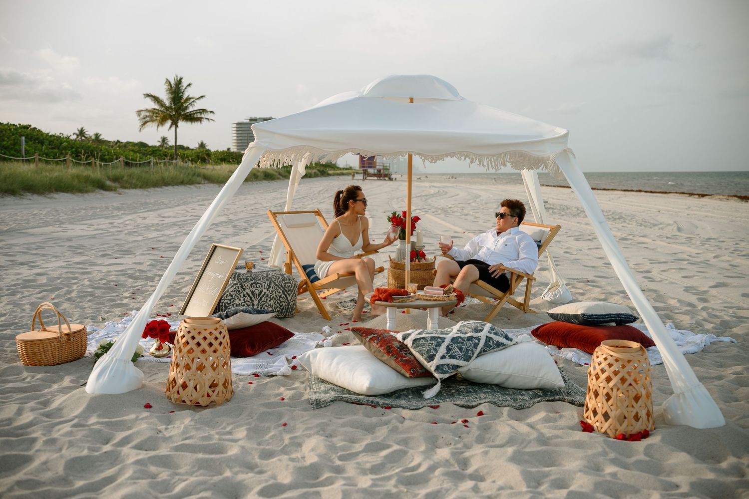 Couple enjoying a romantic beach picnic under a white canopy with lanterns, pillows, and wine on sandy shore.