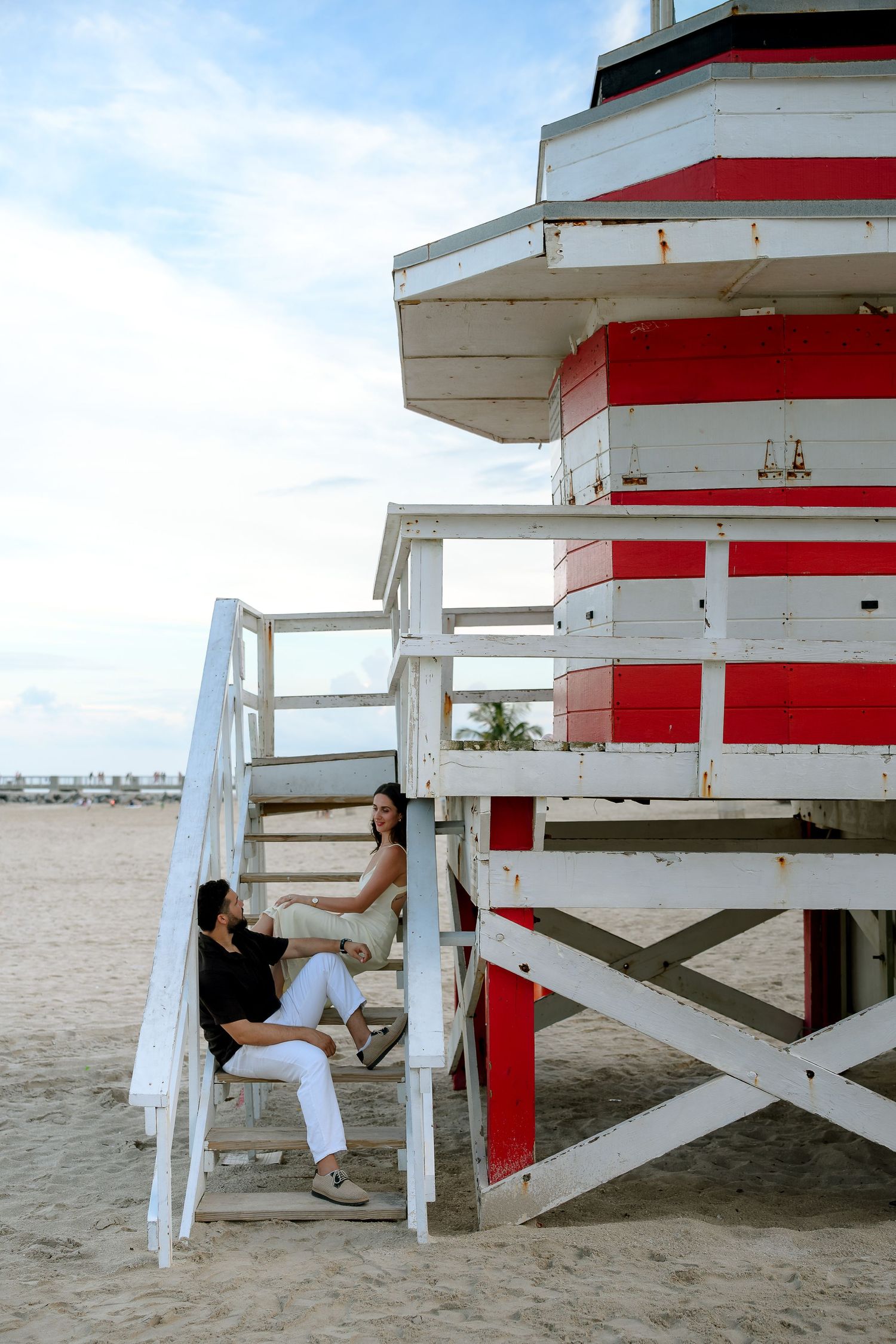 Two women sitting and chatting on the stairs of a red and white lifeguard tower on a sandy Miami Beach.