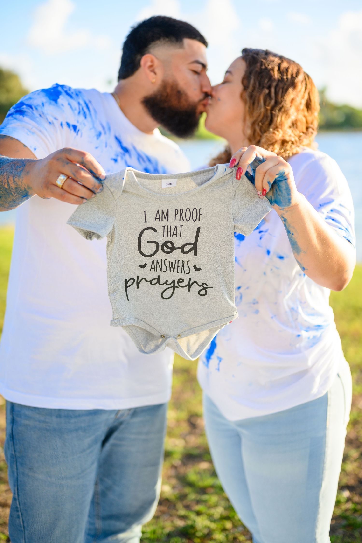 Couple kissing outdoors holding a baby onesie that reads I Am Proof That God Answers Prayers at a gender reveal.