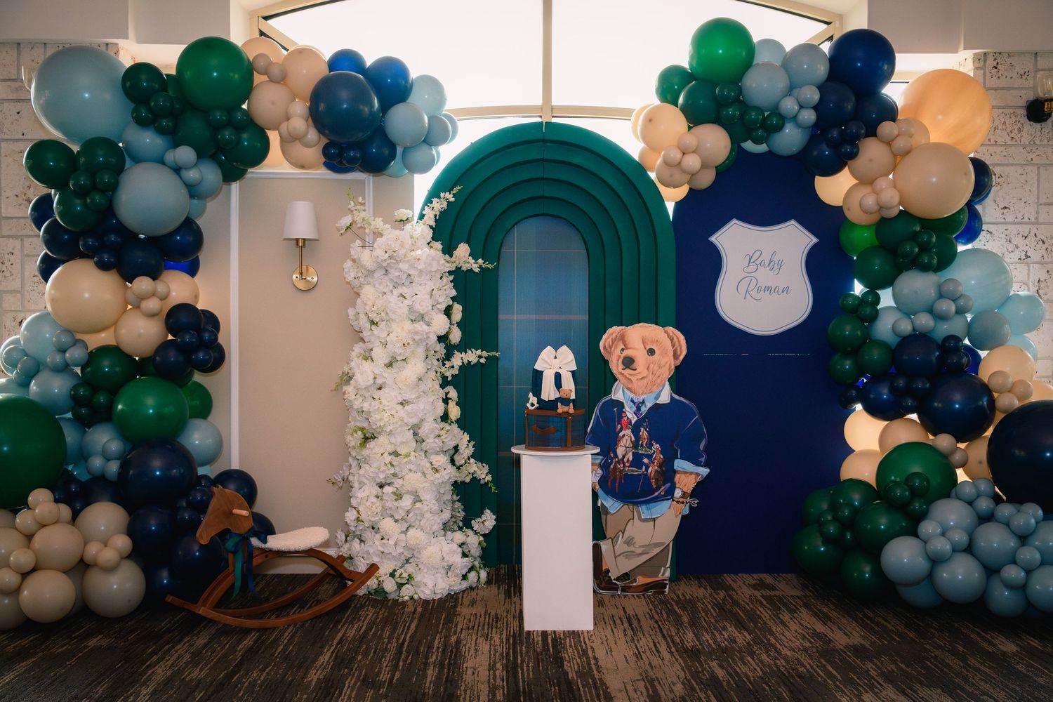 Elegant baby shower backdrop with green and navy balloon arch, teddy bear cutout, and floral decorations.