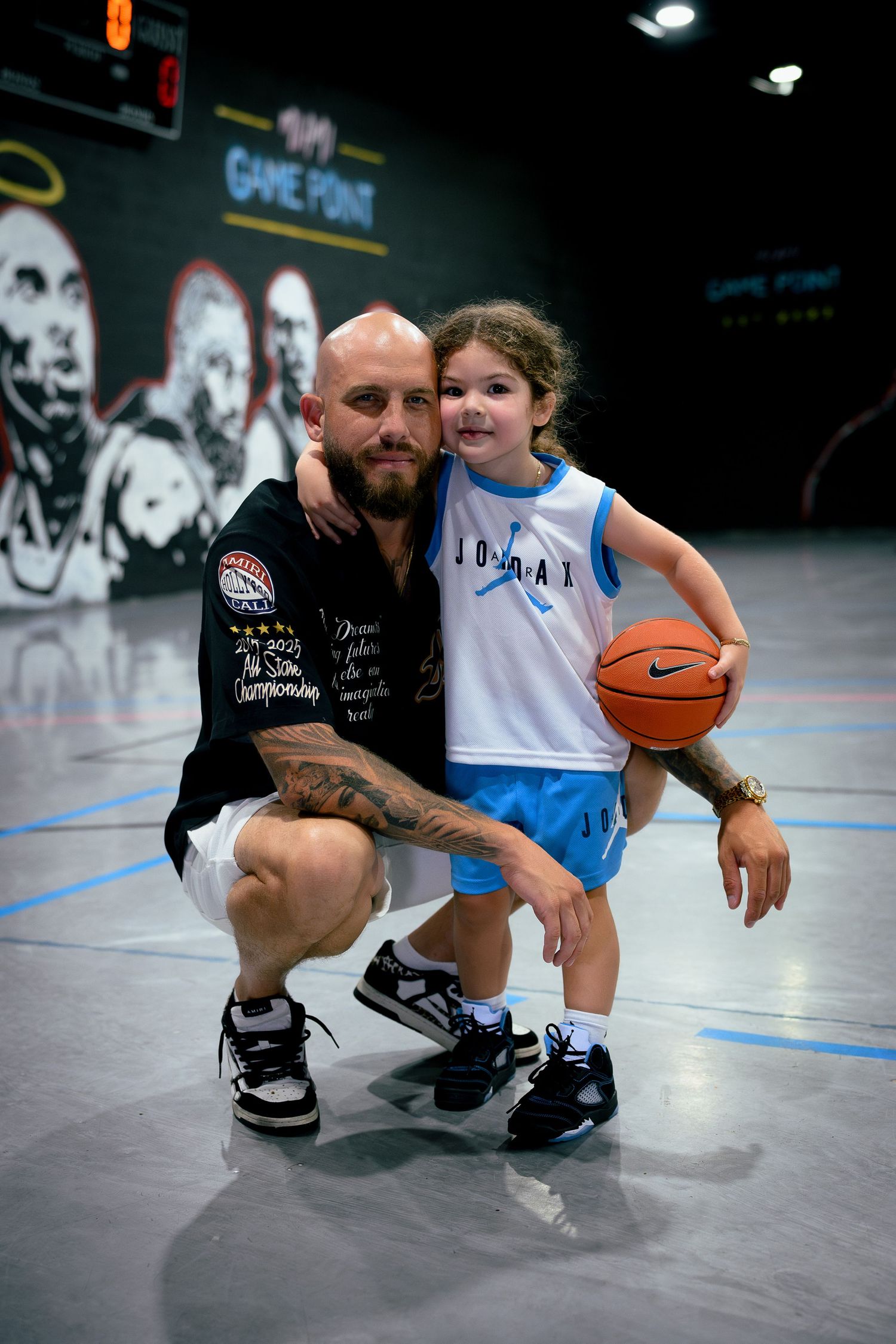 A bald tattooed man crouches beside a young girl holding a basketball on an indoor court at Game Point facility.