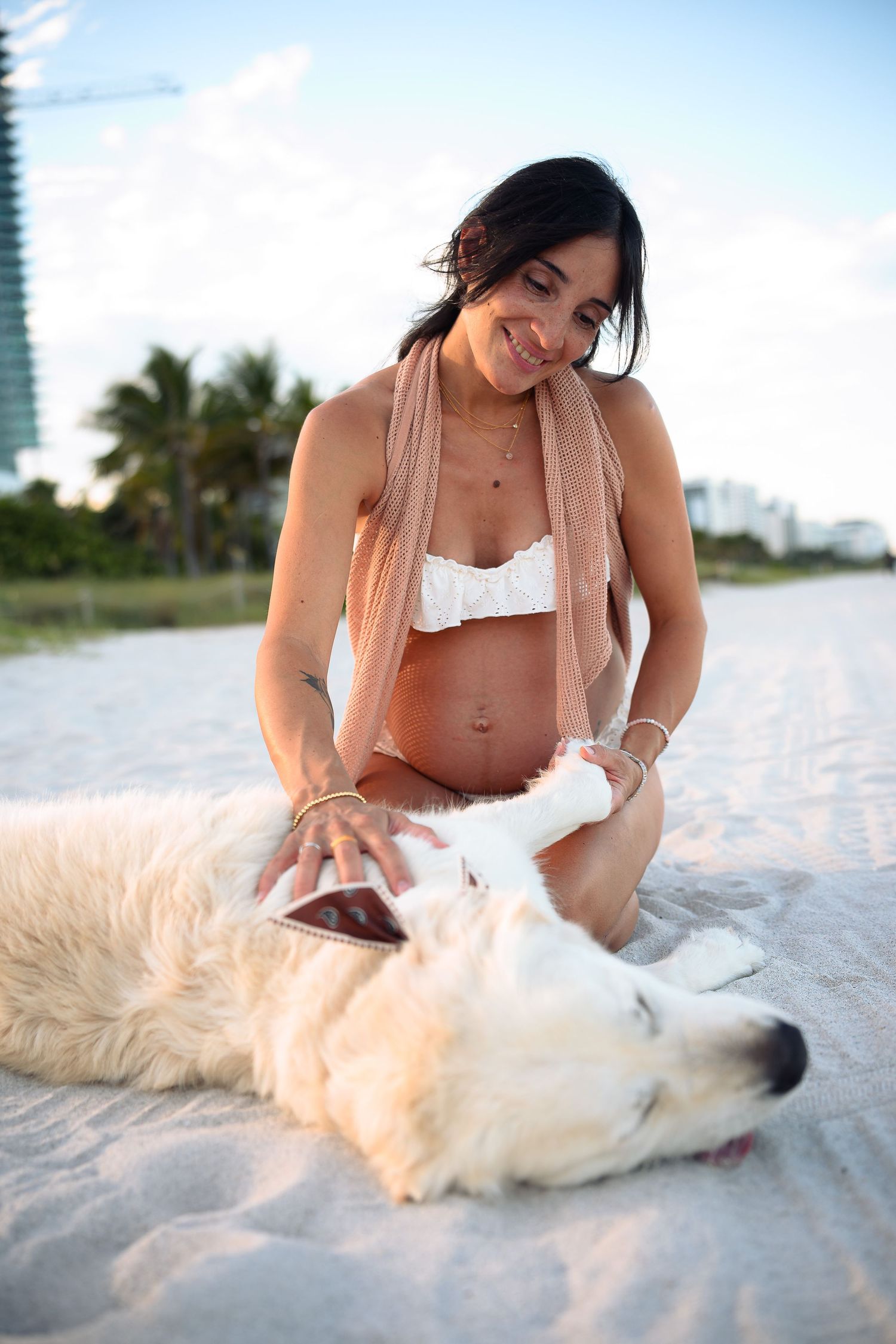 Pregnant woman in white bikini petting a white dog on a sandy beach with palm trees in the background.