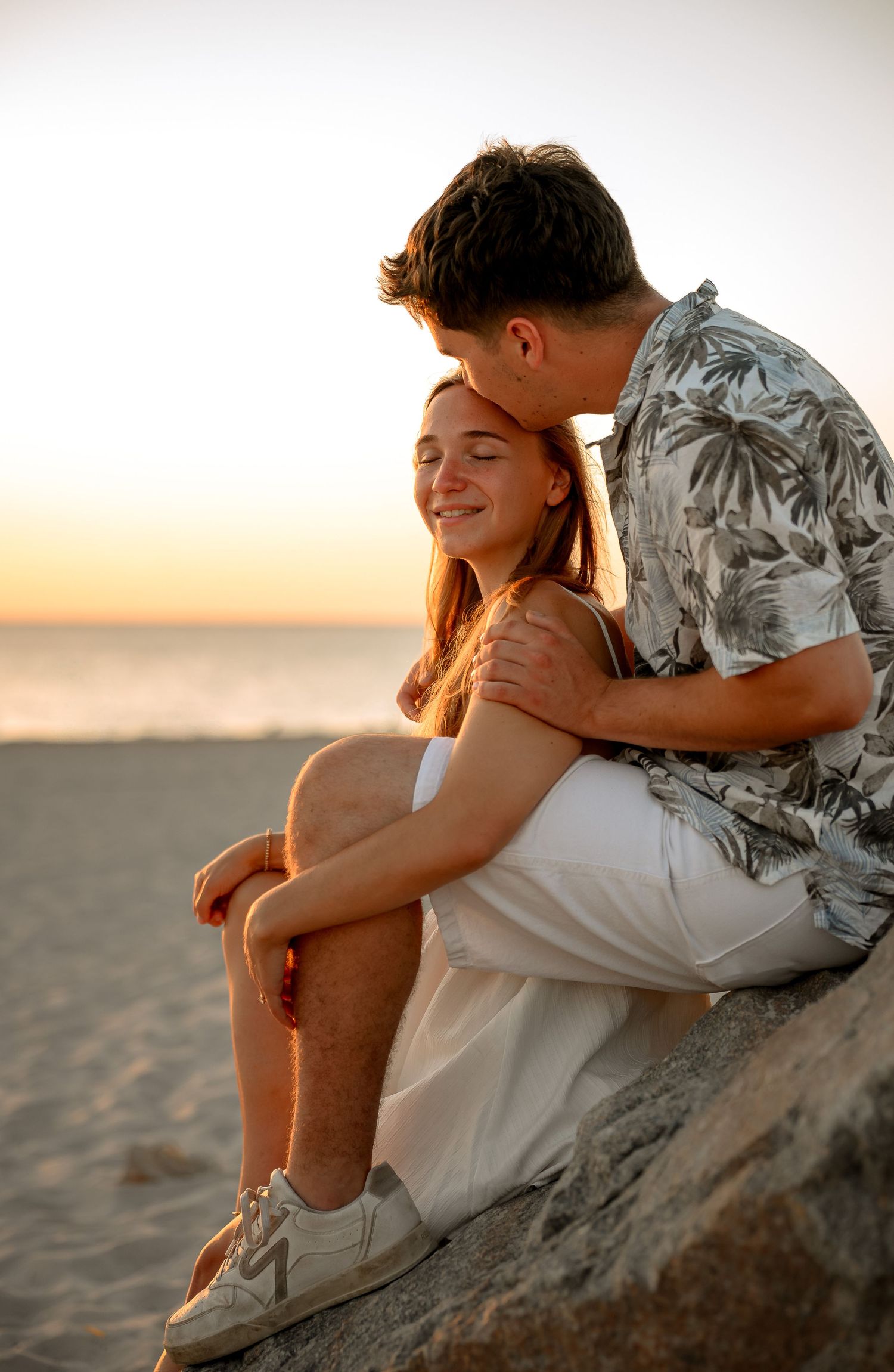 Couple sharing a romantic sunset moment on a beach, man kissing woman's head while she smiles warmly.