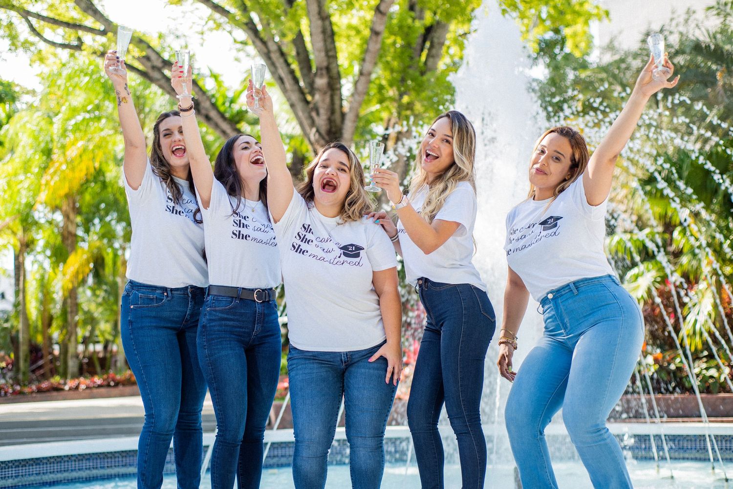 Five women in white graduation tees and jeans celebrate joyfully near a fountain outdoors.