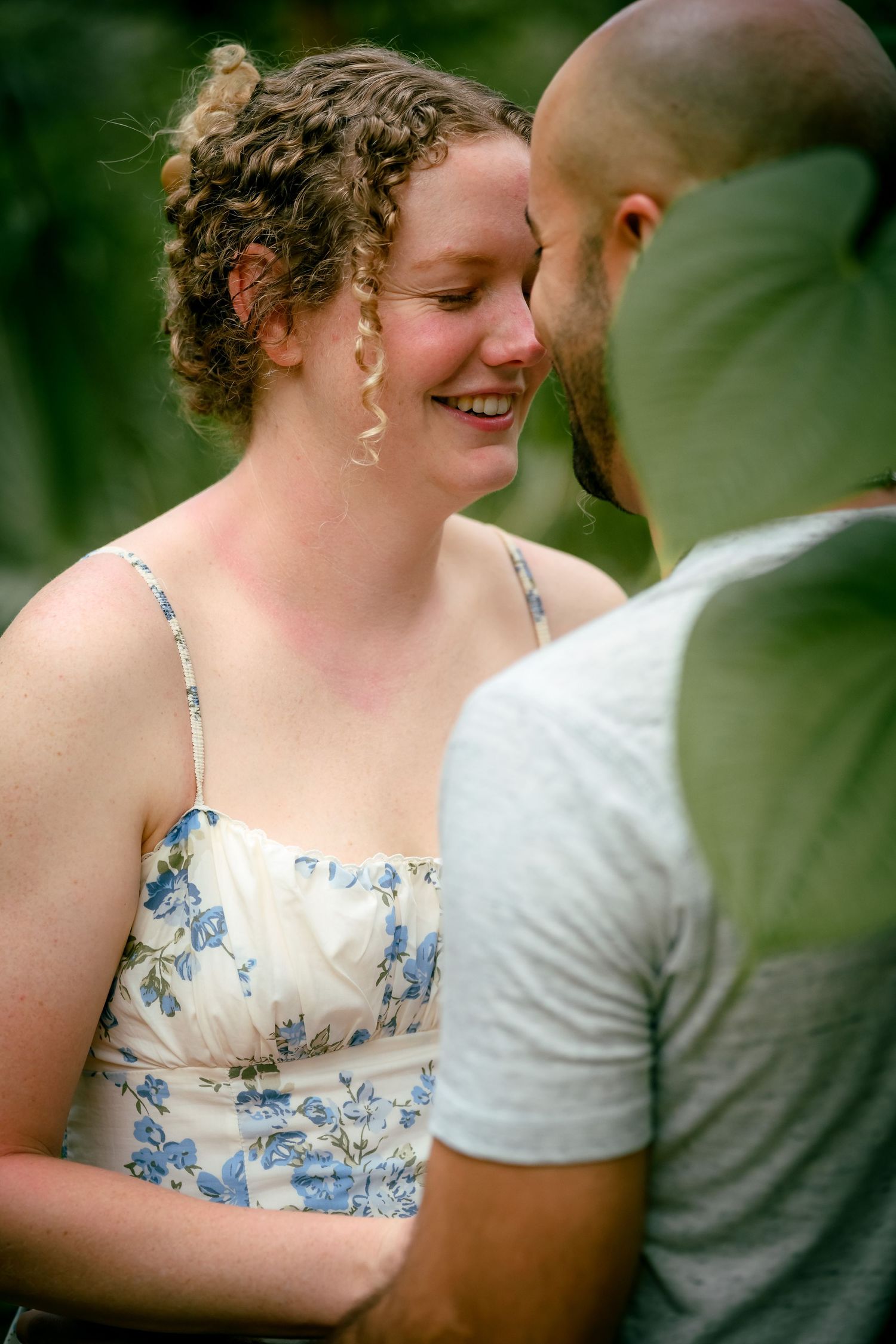 A smiling woman in a floral sundress shares a close romantic moment with a bald man outdoors.