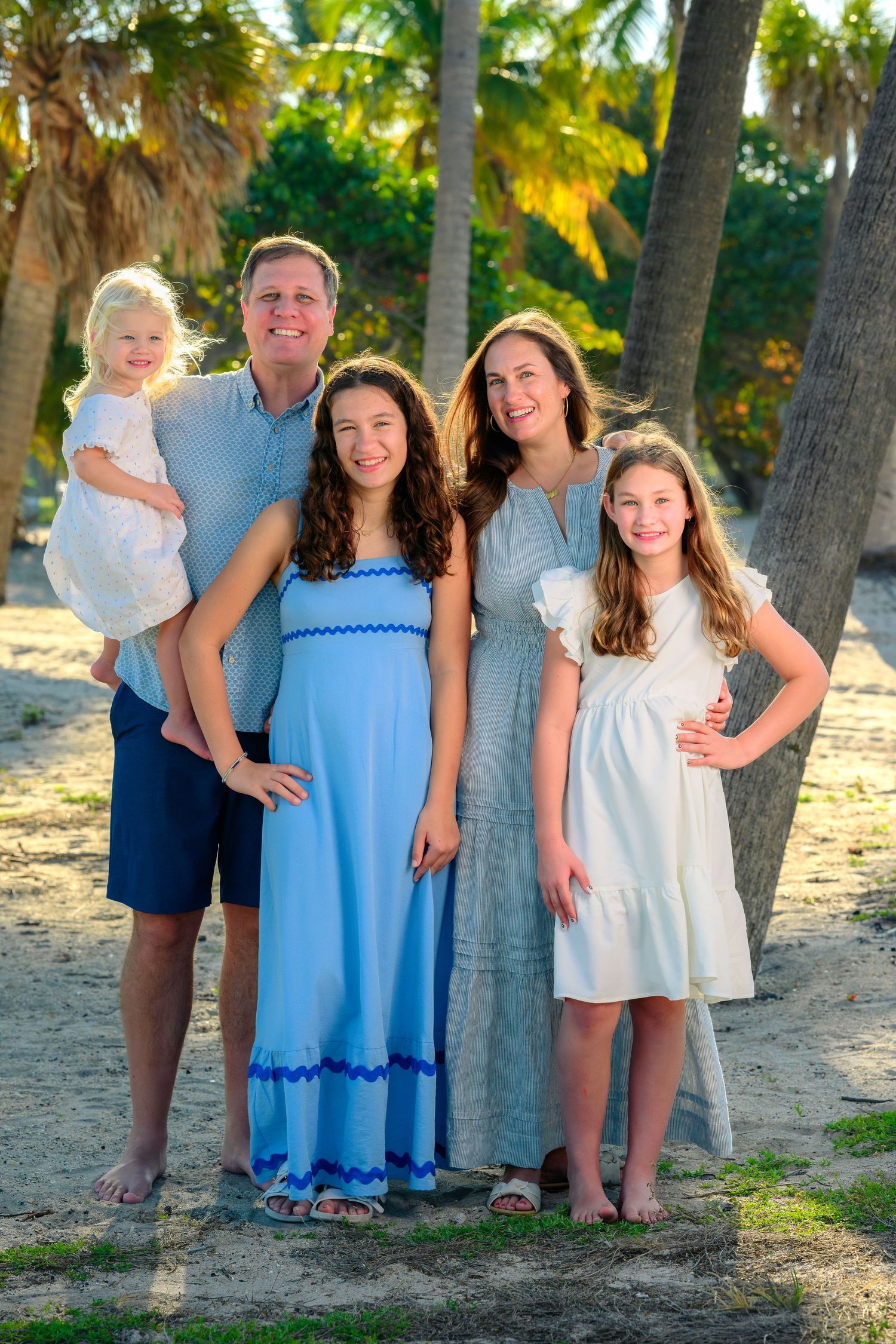 A family of five smiles together on a sandy beach surrounded by tall palm trees.