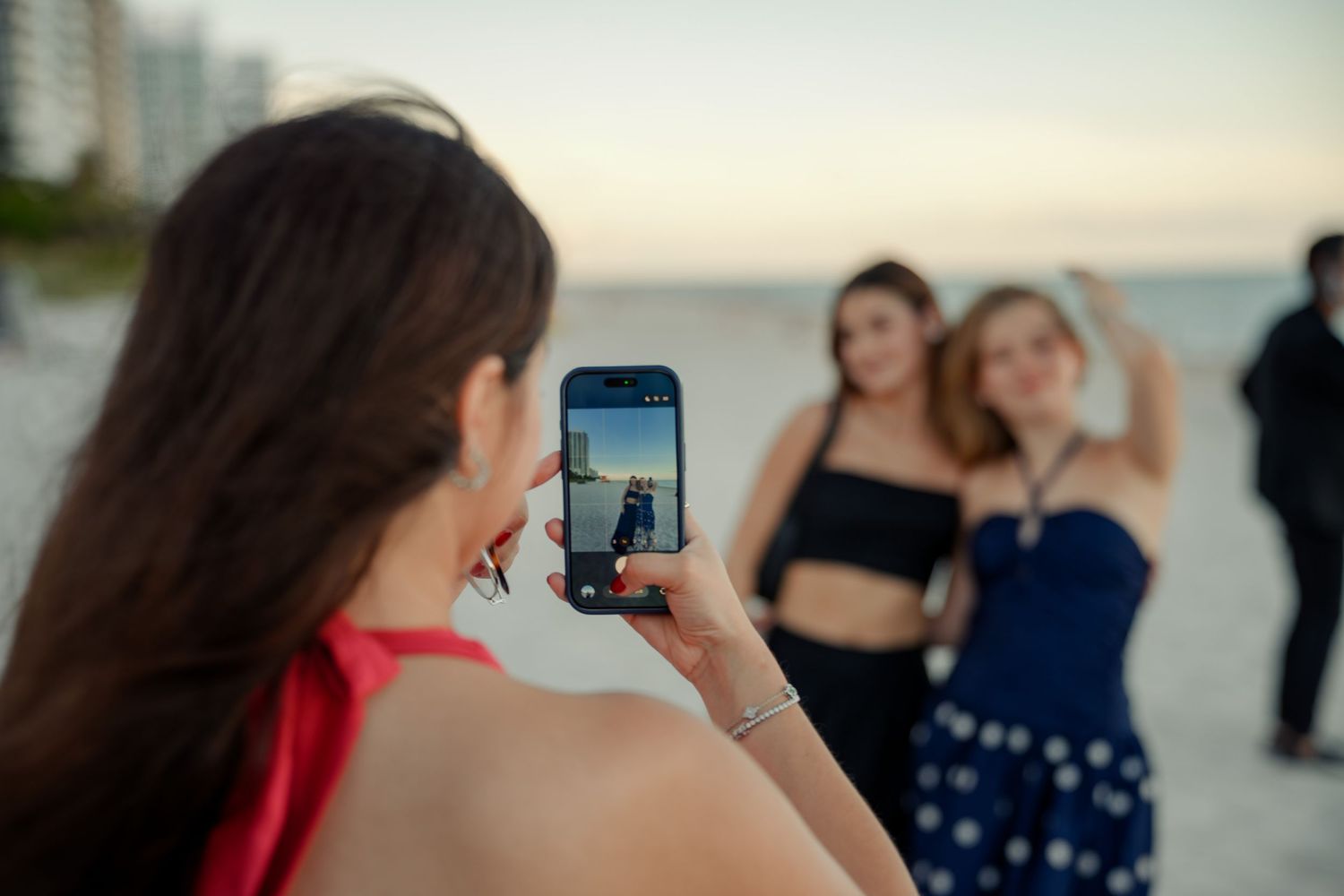 Woman photographing two friends posing on a sandy beach at dusk using a smartphone.