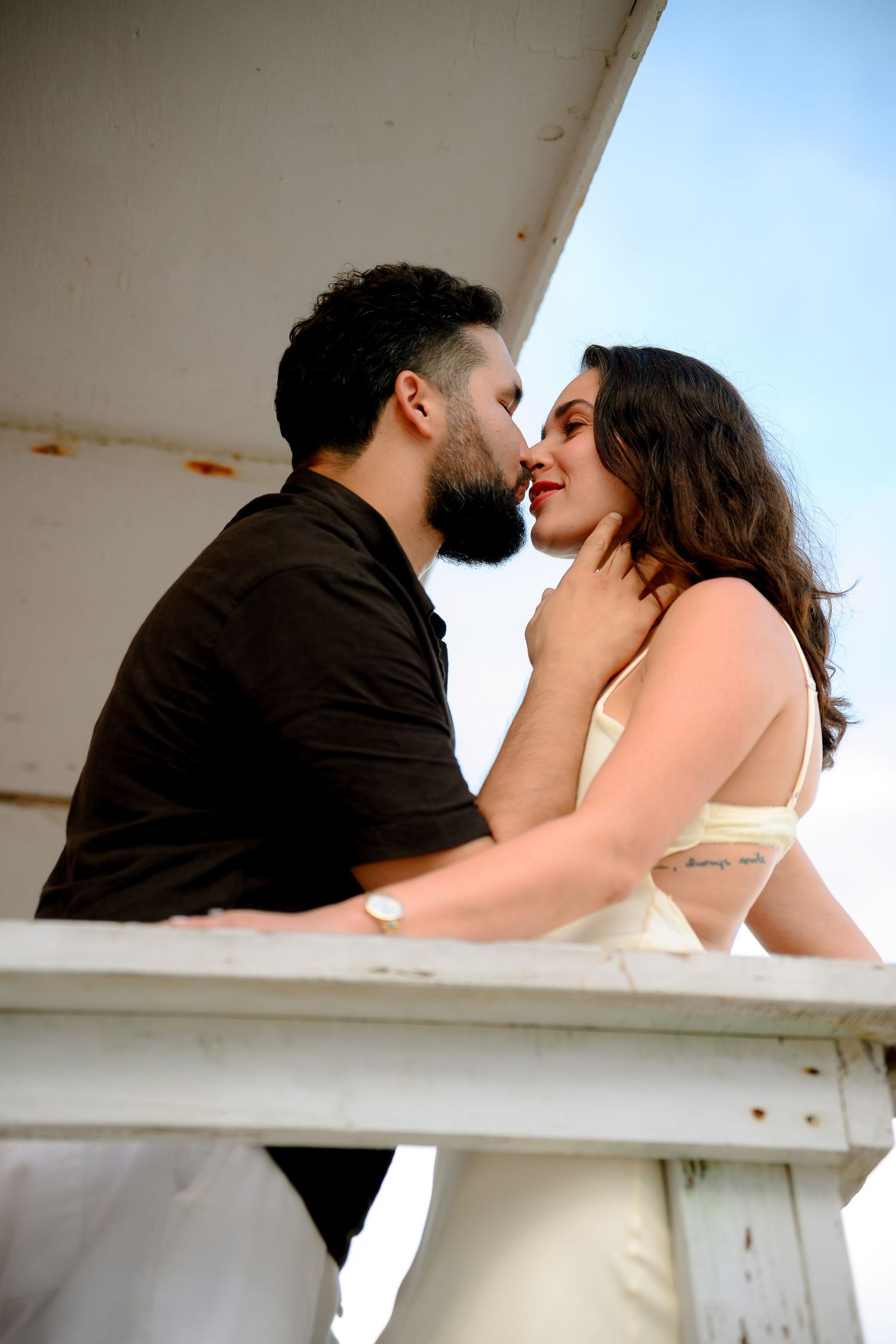 A romantic couple shares an intimate moment on a white balcony, the bearded man tenderly holding the woman's face.